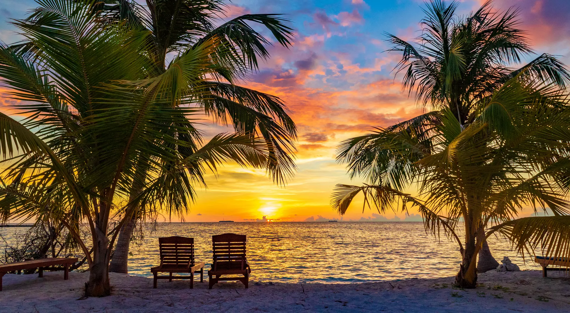 Tropical beach at sunset with palm trees and two chairs facing the ocean, vibrant sky in shades of orange and blue.