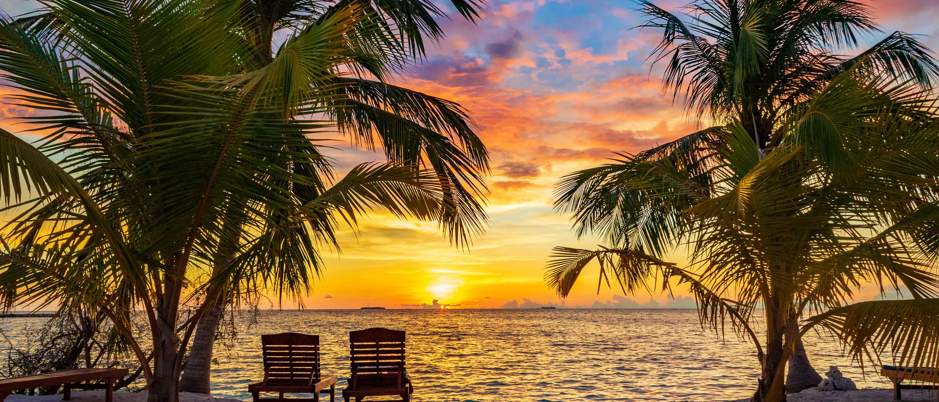 Tropical beach at sunset with palm trees and two chairs facing the ocean, vibrant sky in shades of orange and blue.