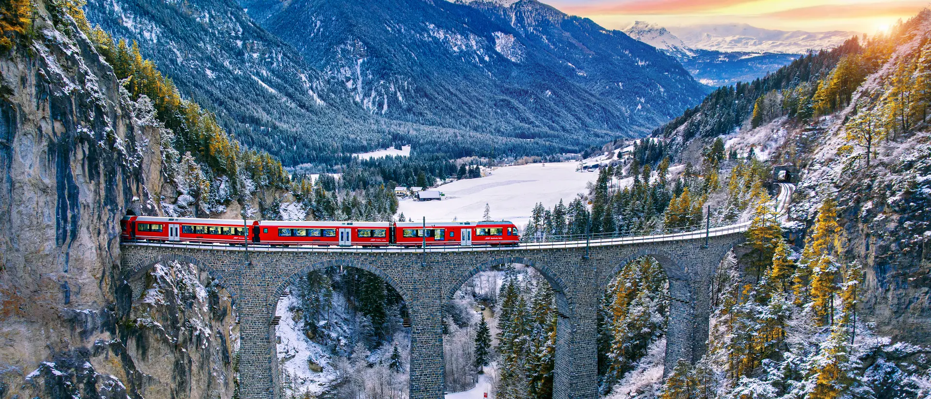 A red train crosses a stone bridge over a valley, surrounded by snow-capped mountains and evergreen trees at sunset.