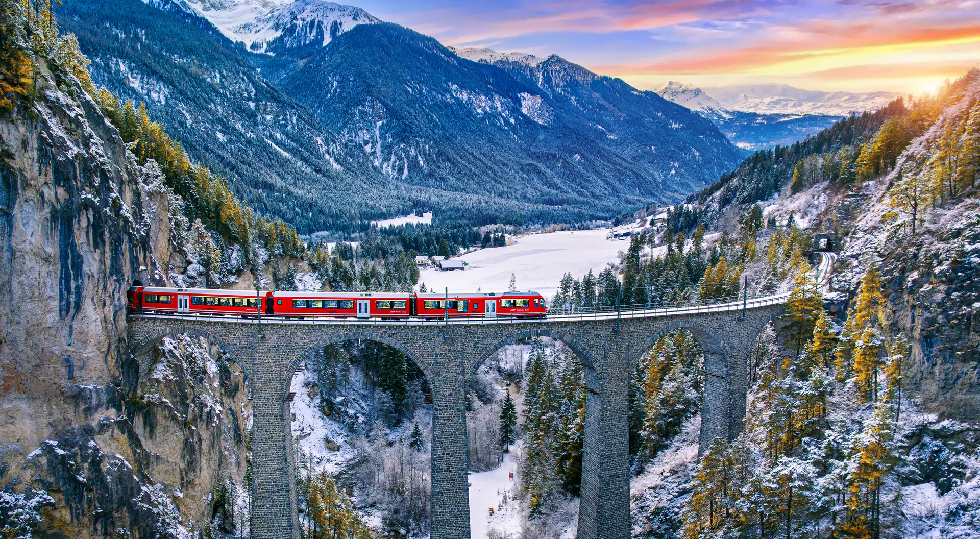 A red train crosses a stone bridge over a valley, surrounded by snow-capped mountains and evergreen trees at sunset.