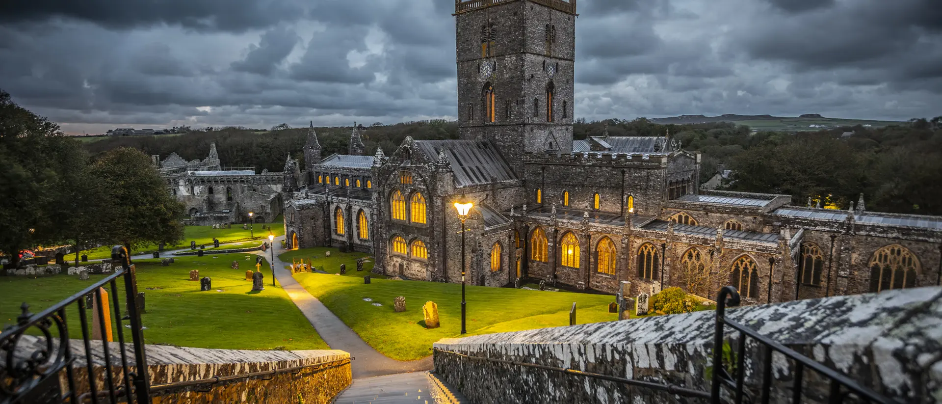 Historic stone church with a tall tower, surrounded by trees and glowing lights, under a dramatic cloudy sky.