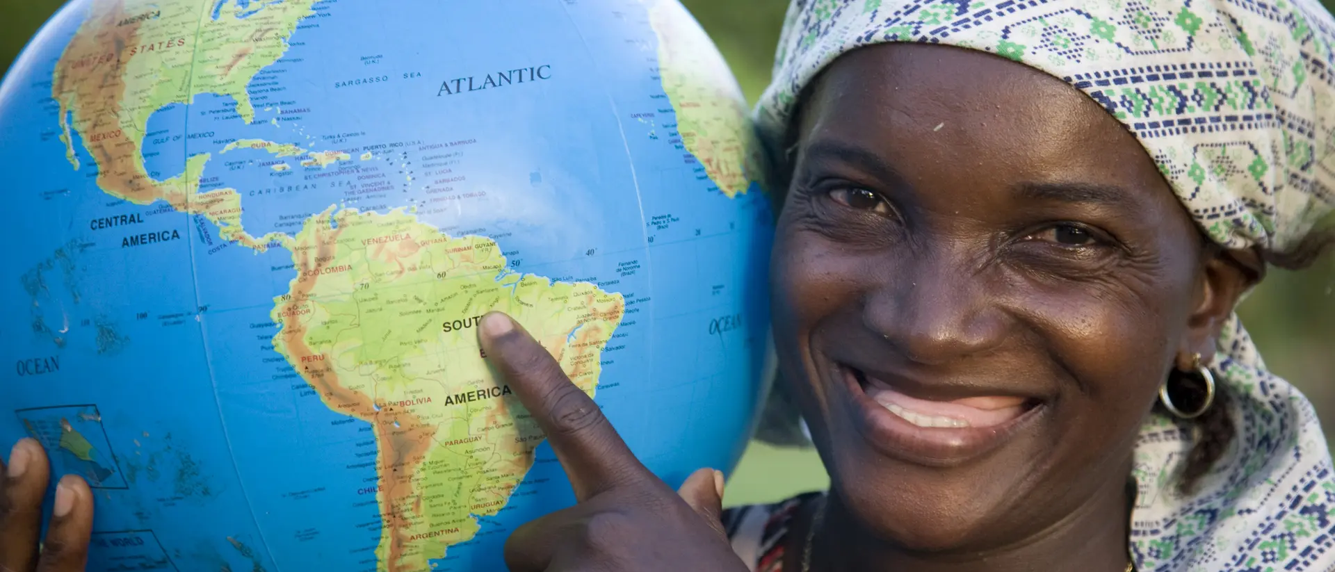 Smiling woman pointing at a globe, highlighting South America, against a natural backdrop.