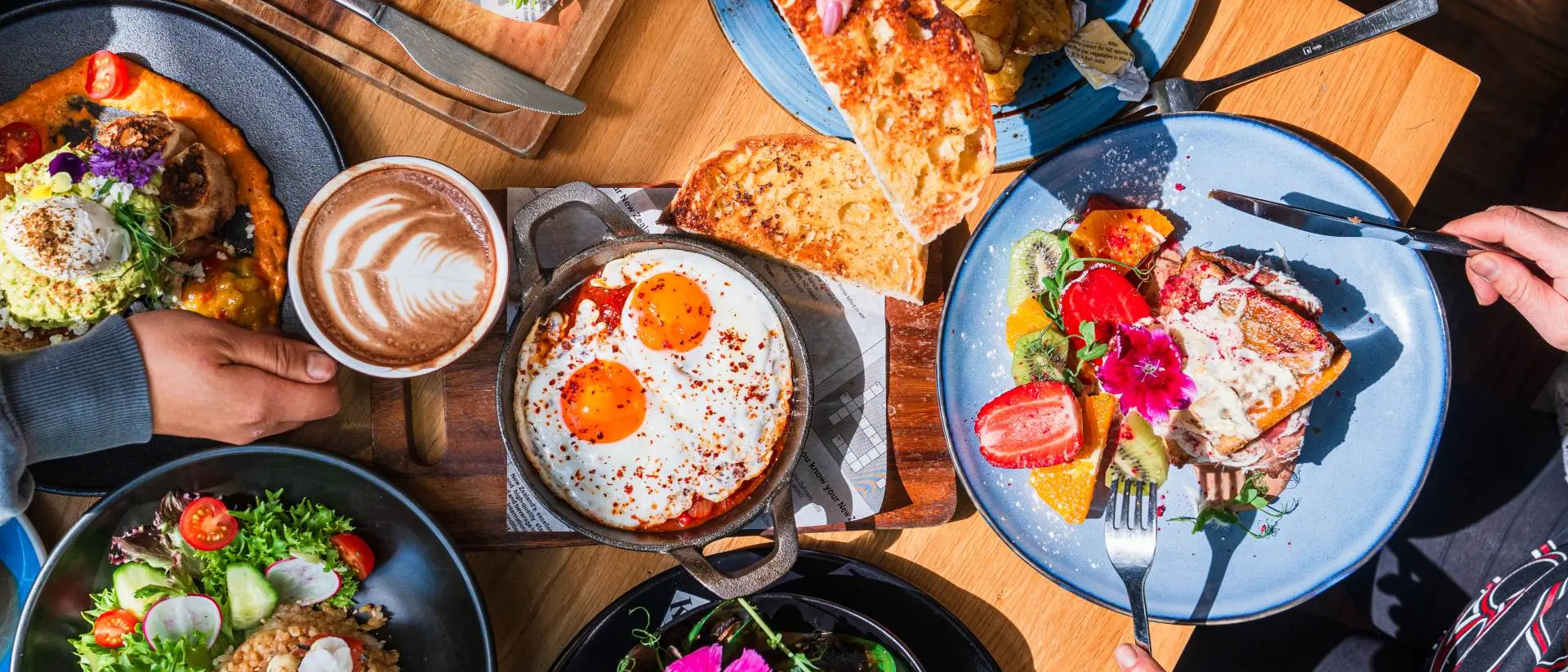 A top view of a diverse brunch spread with dishes, drinks, and hands sharing food around a wooden table.