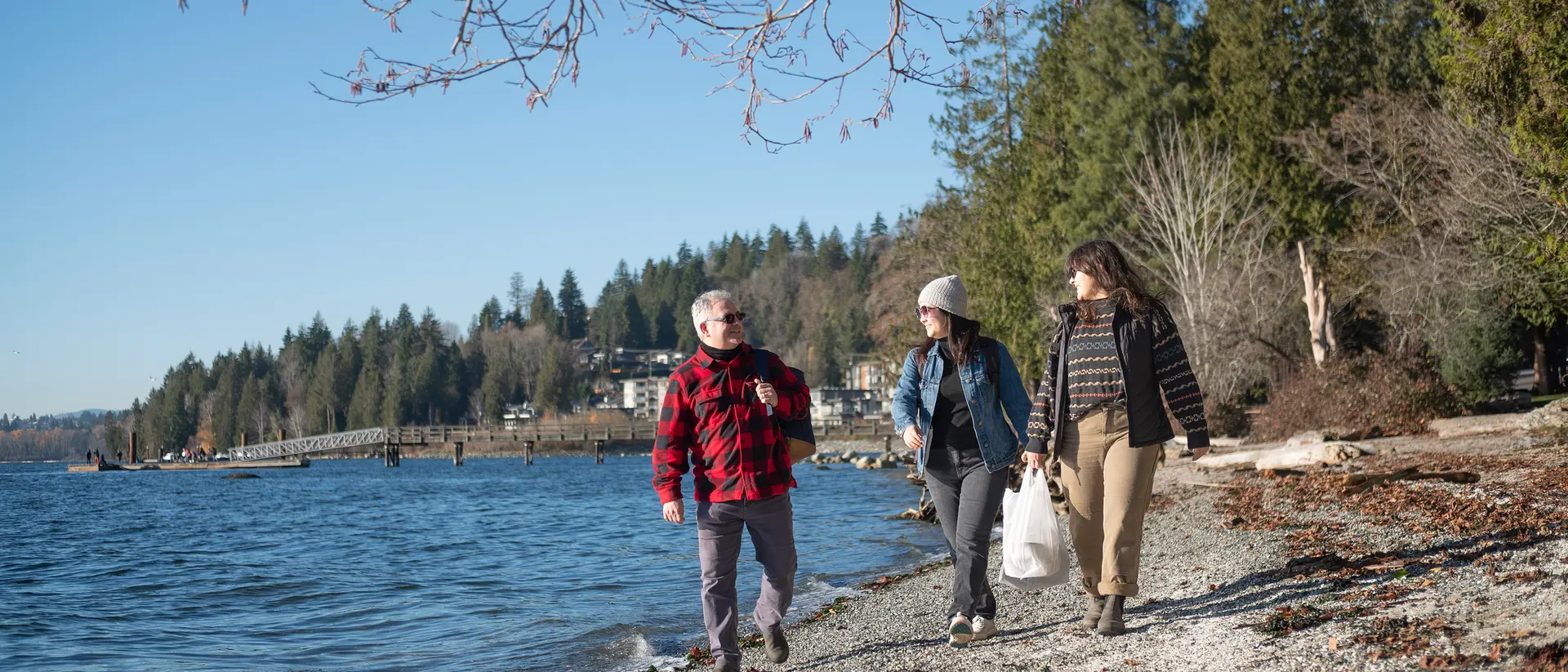 Three people walk along a beach by a lake, surrounded by trees and a clear blue sky.