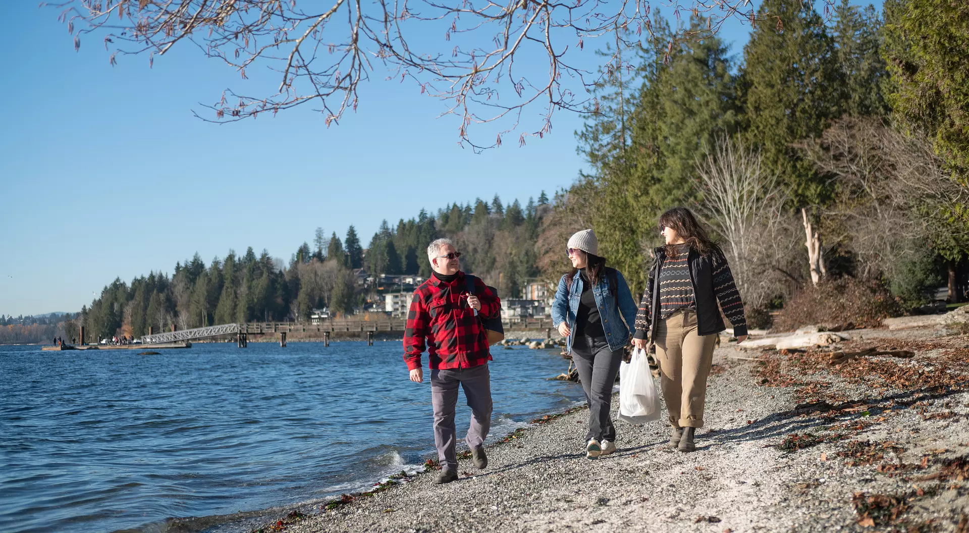 Three people walk along a beach by a lake, surrounded by trees and a clear blue sky.