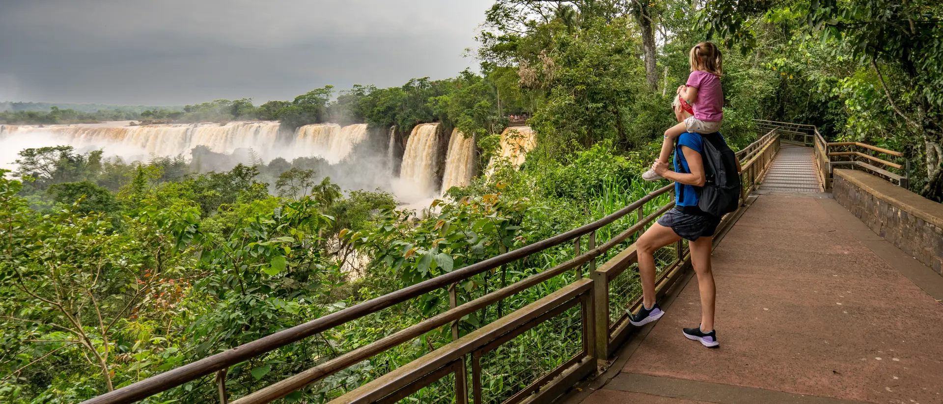 A person with a child on their back stands on a path overlooking a lush landscape and waterfalls in the distance.
