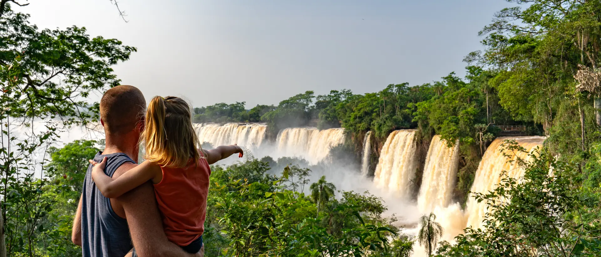 A parent holds a child while pointing at a cascading waterfall surrounded by lush greenery.