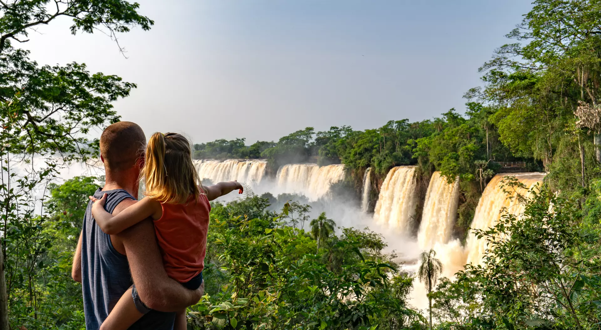 A parent holds a child while pointing at a cascading waterfall surrounded by lush greenery.