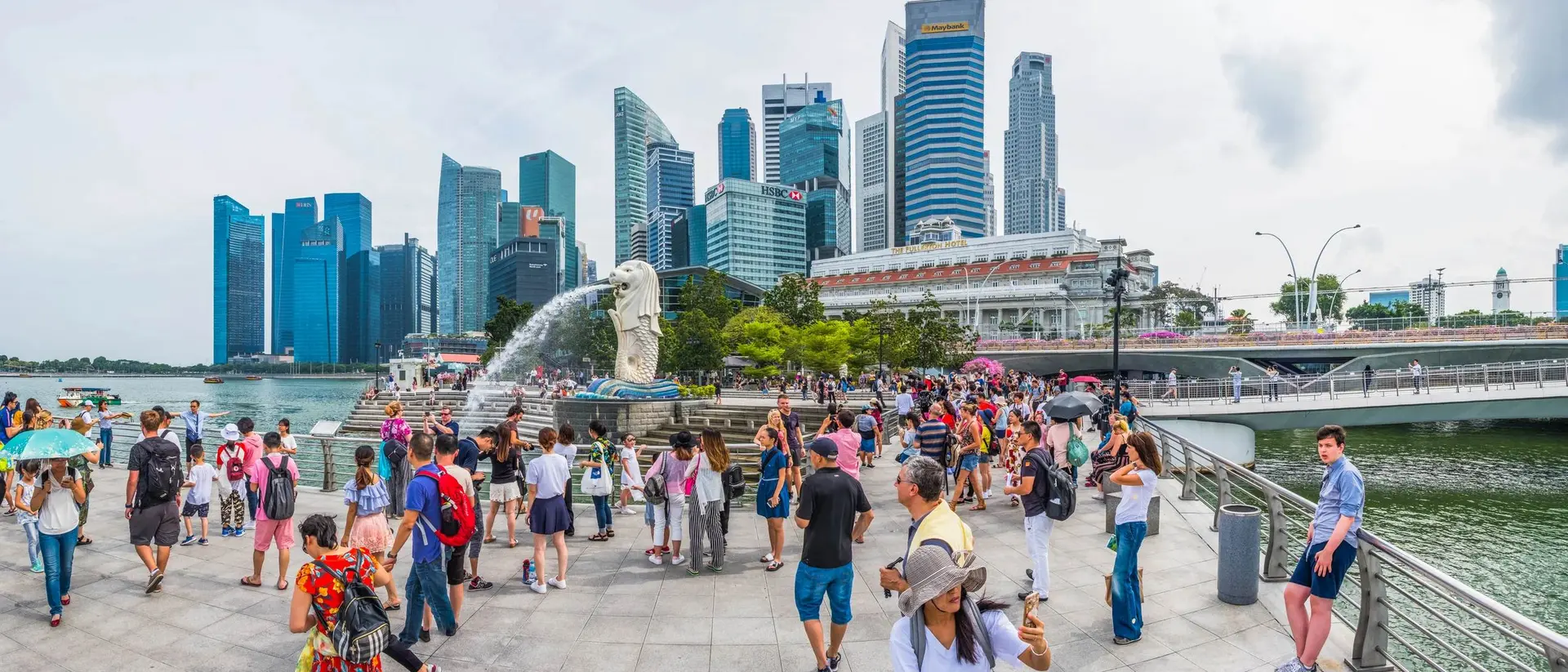 Crowd gathered by the Merlion statue with Singapore's skyline in the background, featuring modern skyscrapers.