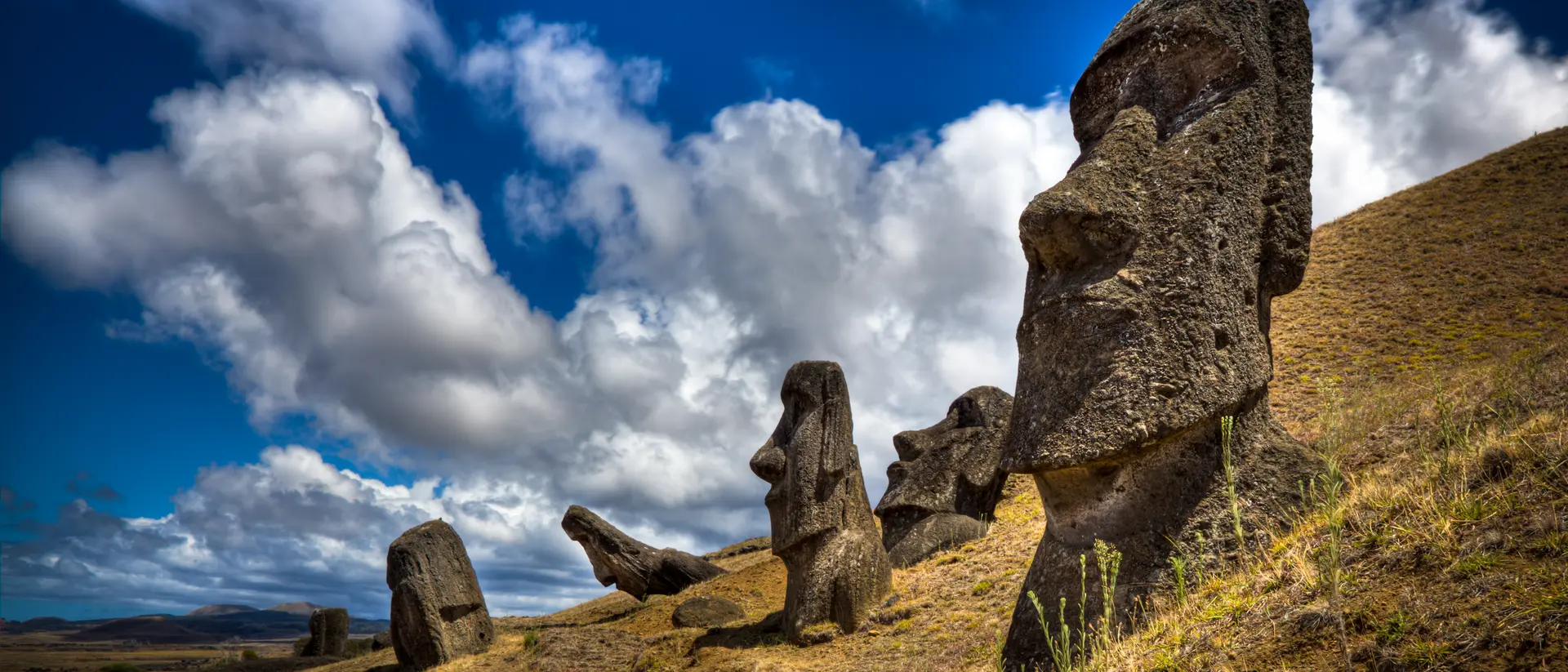 One Moais is in the foreground and four in the background. The sky is mostly sunny with clouds.