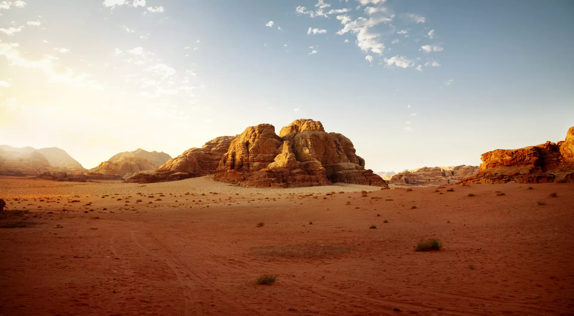 Desert landscape with rock formations under a clear sky, highlighted by warm sunlight and scattered clouds.