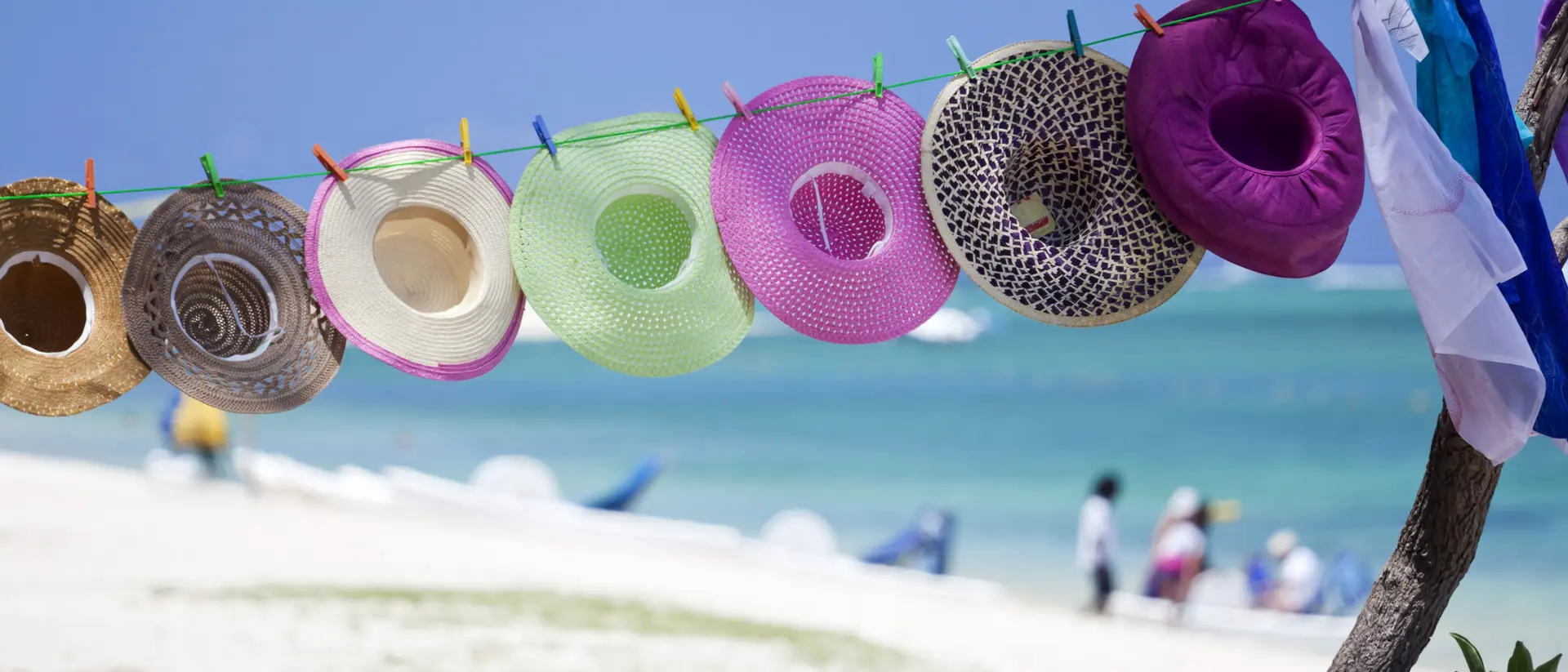 Colorful hats hang on a line with a beach and ocean in the background, accompanied by people enjoying the scenery.