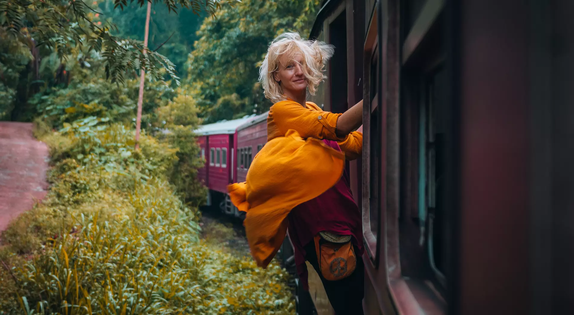 A person in a bright yellow dress leans out of a train window, surrounded by lush greenery and a second train in the background.