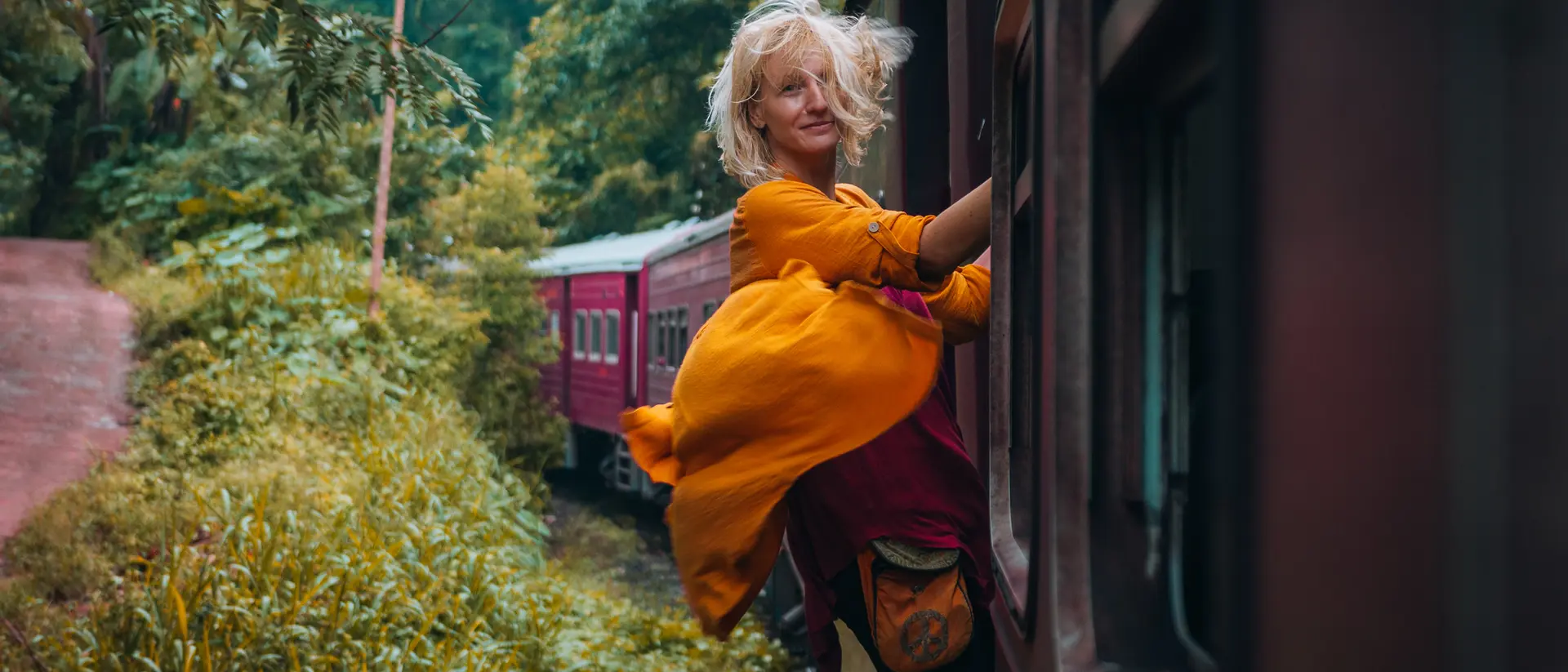 A person in a bright yellow dress leans out of a train window, surrounded by lush greenery and a second train in the background.