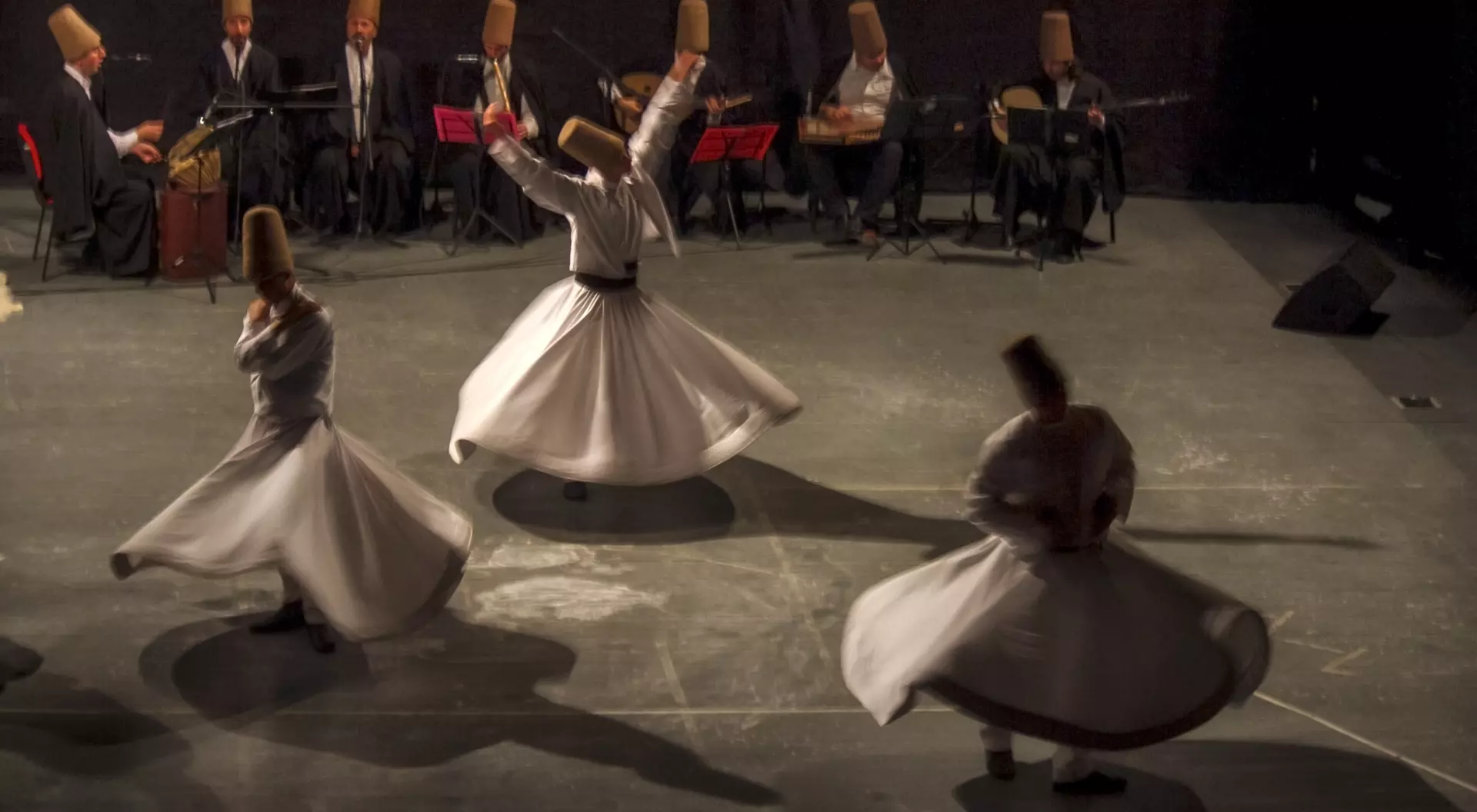 Whirling dervishes perform in front of musicians, all wearing traditional hats, on a dimly lit stage.