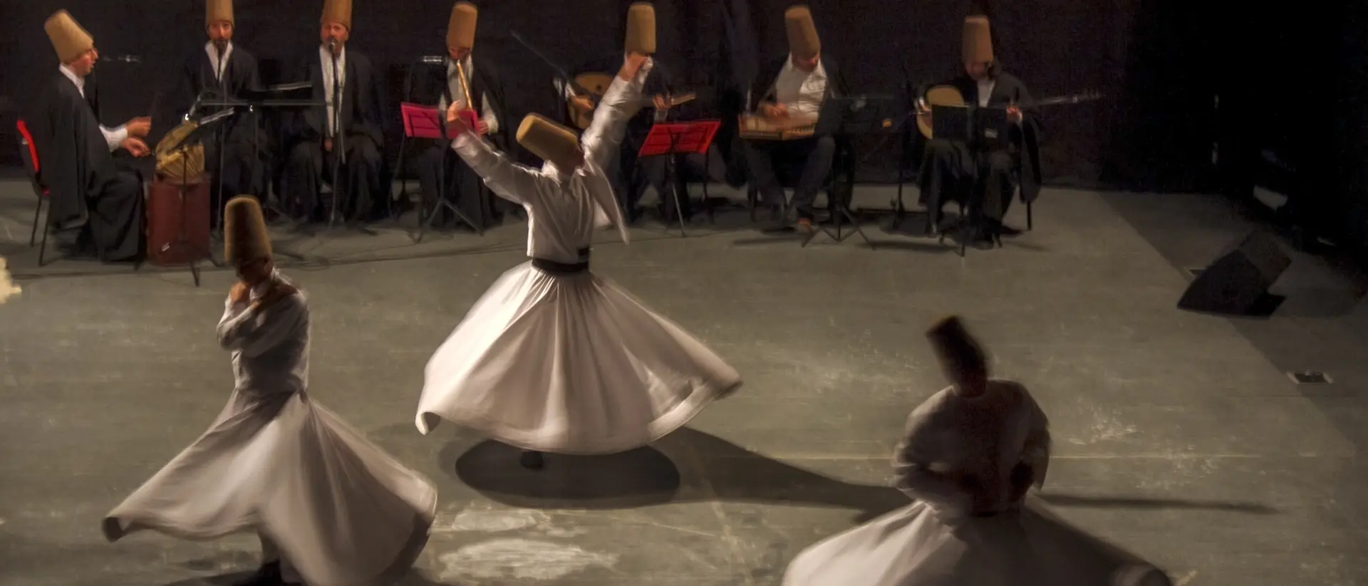 Whirling dervishes perform in front of musicians, all wearing traditional hats, on a dimly lit stage.