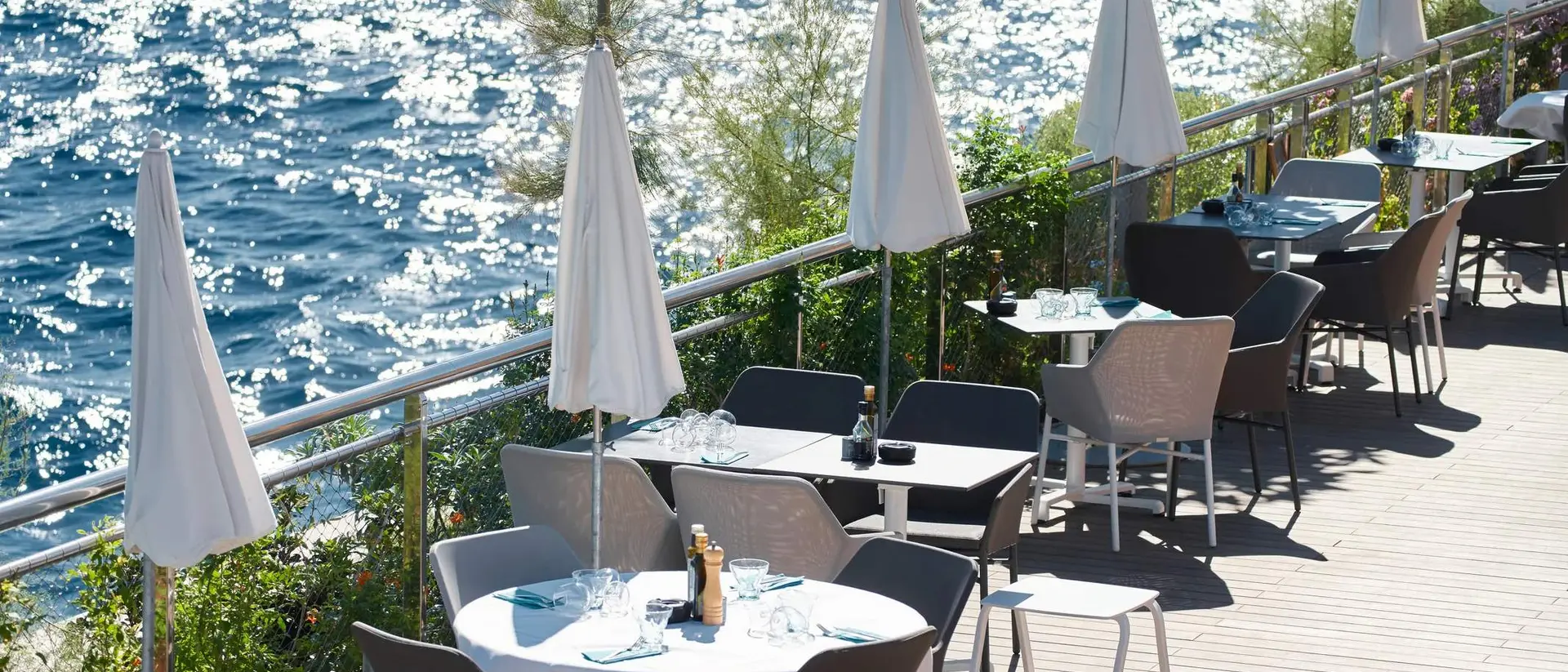 Outdoor restaurant patio with tables and umbrellas beside a sparkling blue sea.