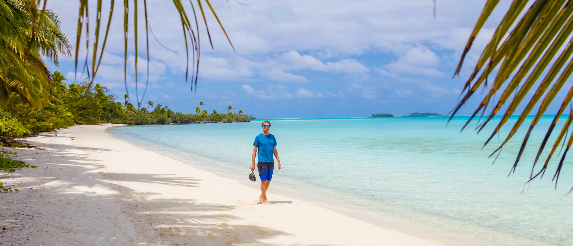 A person walks along a sandy beach with palm trees and clear turquoise water under a partly cloudy sky.