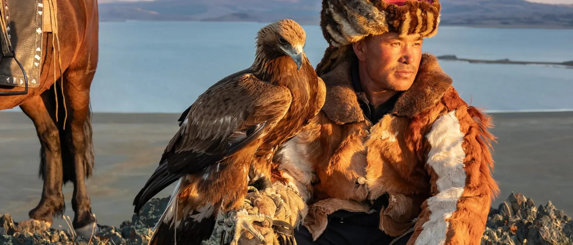 A hunter in traditional attire sits with a golden eagle, overlooking a scenic landscape with a horse nearby.