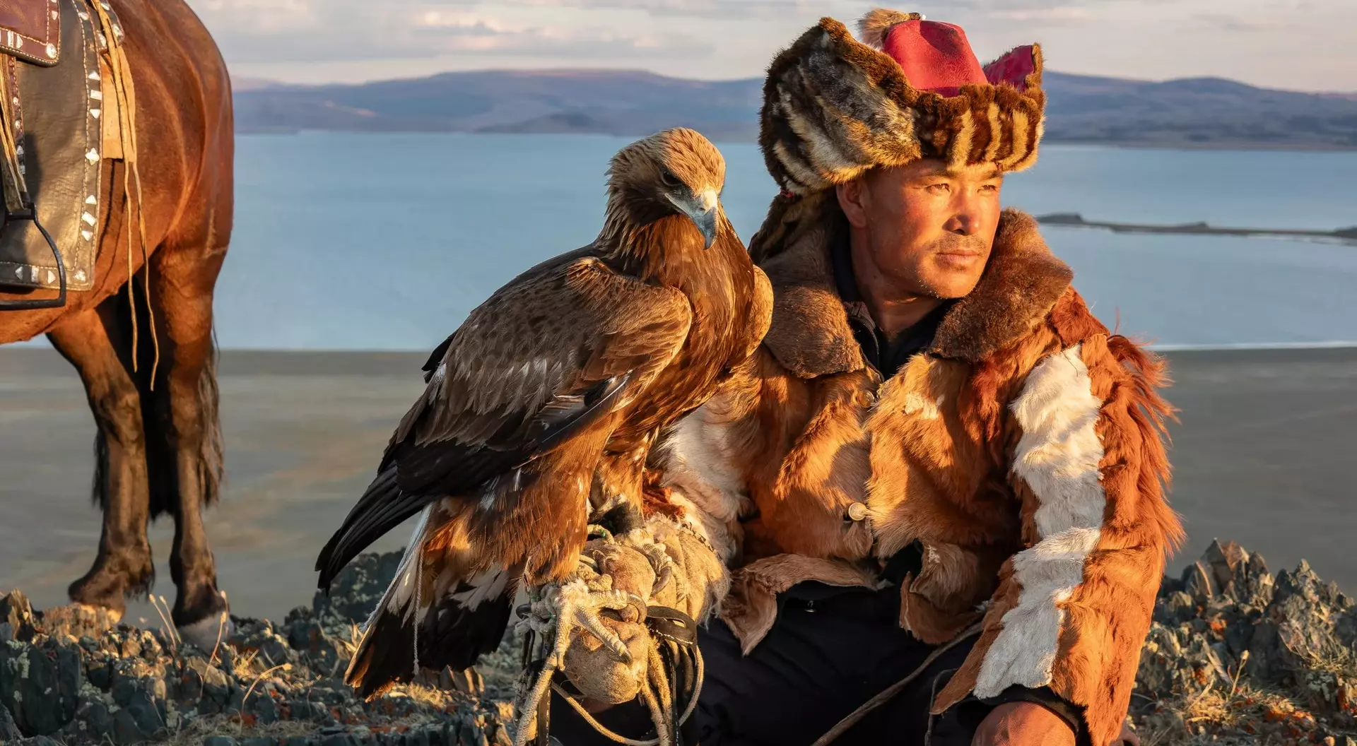 A hunter in traditional attire sits with a golden eagle, overlooking a scenic landscape with a horse nearby.