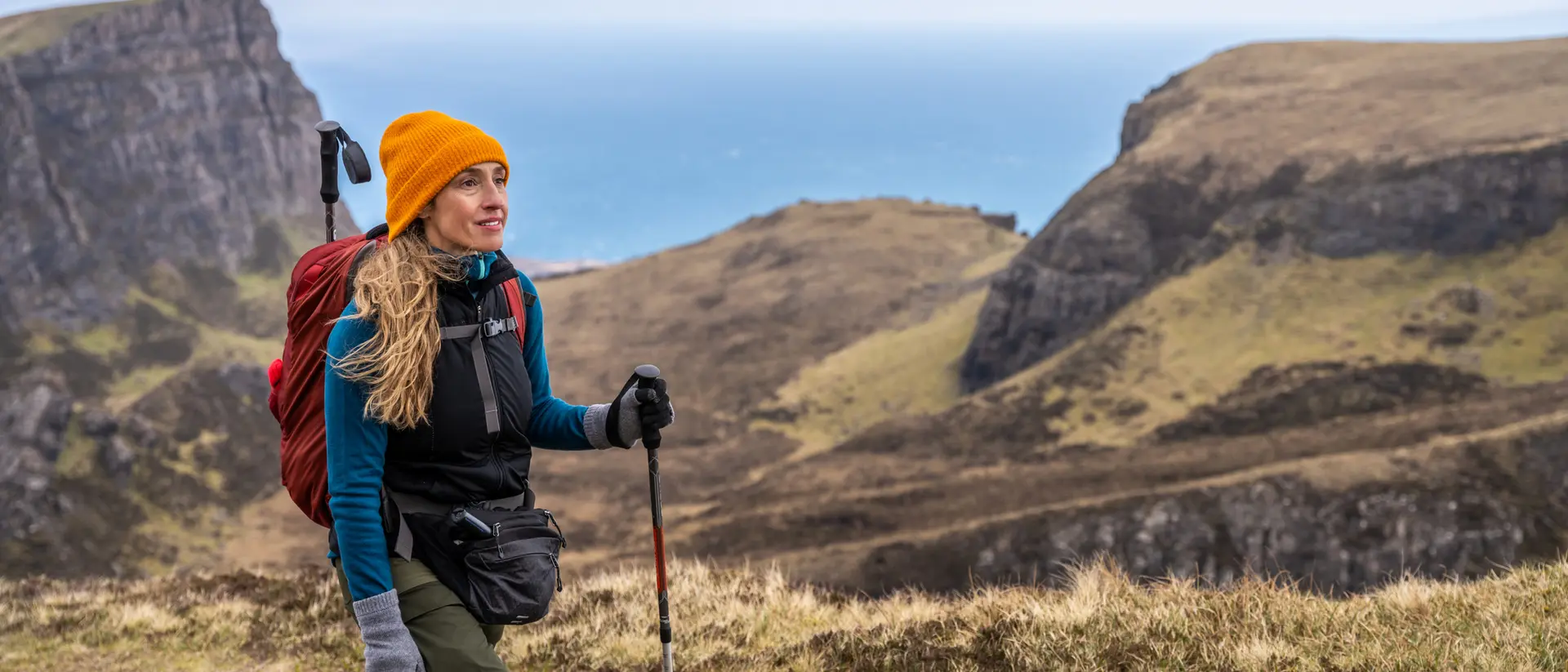 A woman with long hair in a bright orange hat hikes along a coastal path, with cliffs and the ocean in the background.