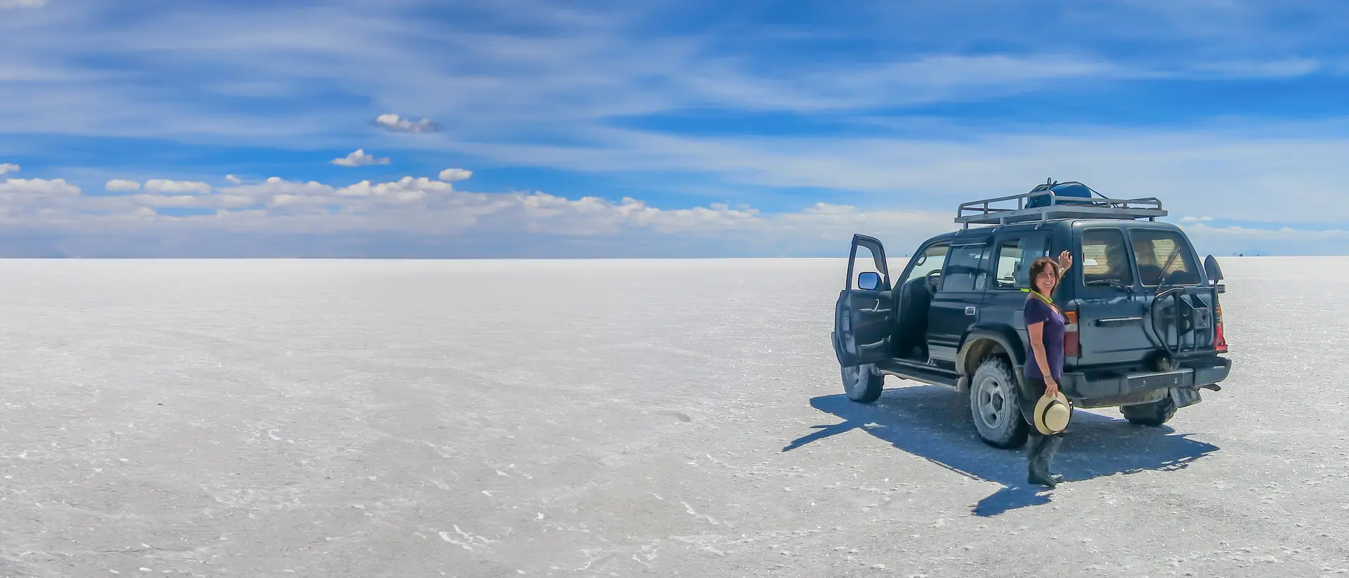 A person stands next to a parked SUV on a vast white salt flat under a blue sky with scattered clouds.