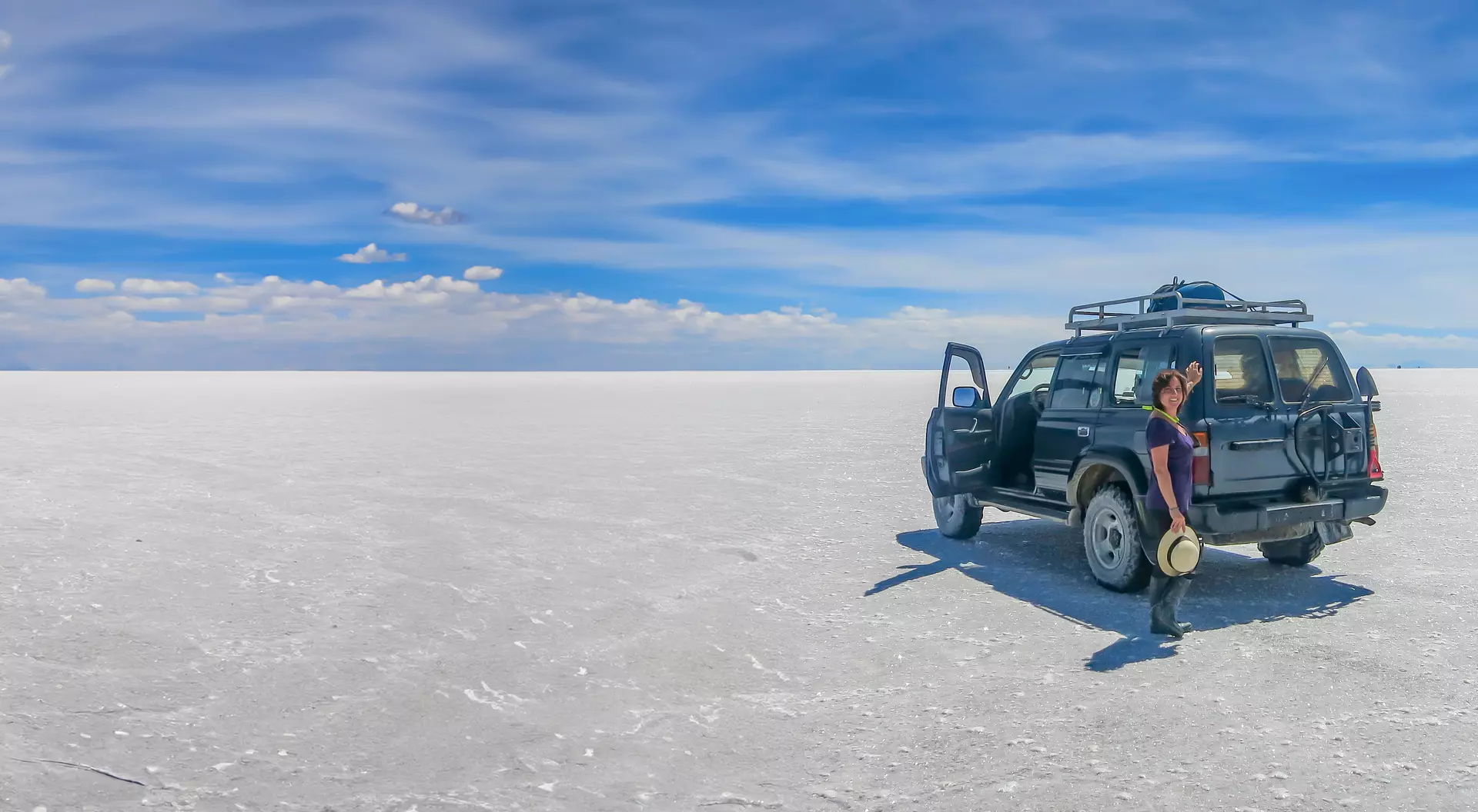 A person stands next to a parked SUV on a vast white salt flat under a blue sky with scattered clouds.