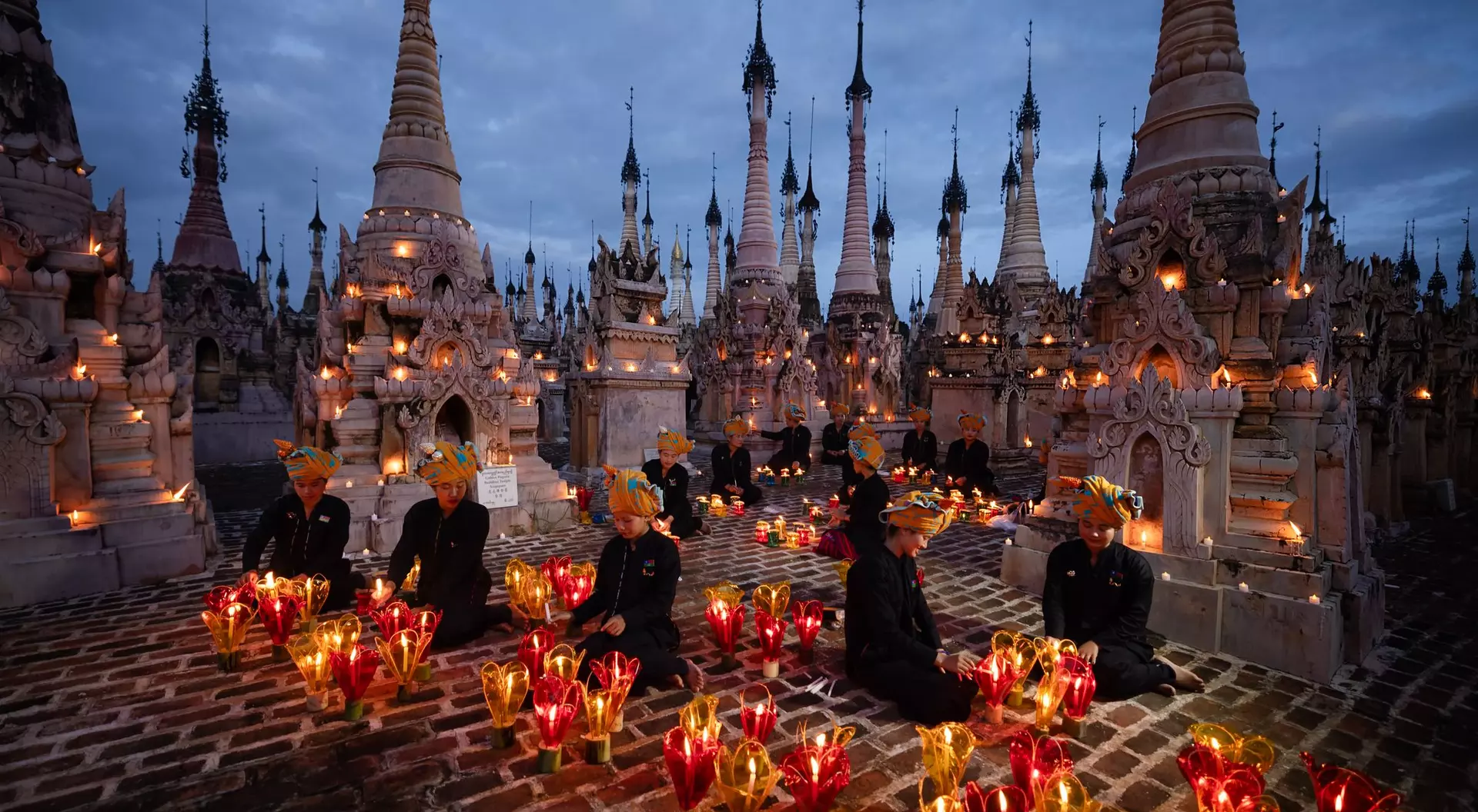 A serene twilight scene with people seated among illuminated pagodas and glowing candles, creating a peaceful atmosphere.