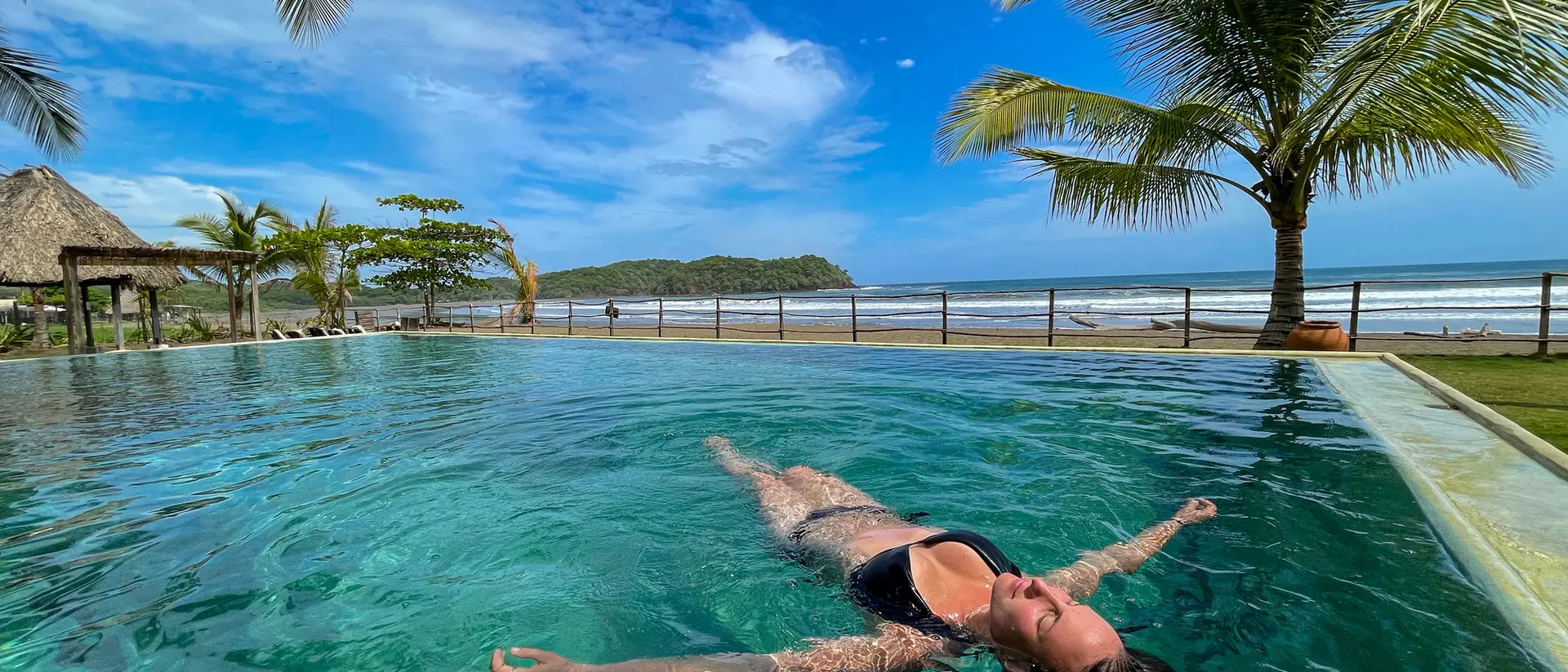 Person floating in a pool with palm trees and ocean in the background under a sunny blue sky.