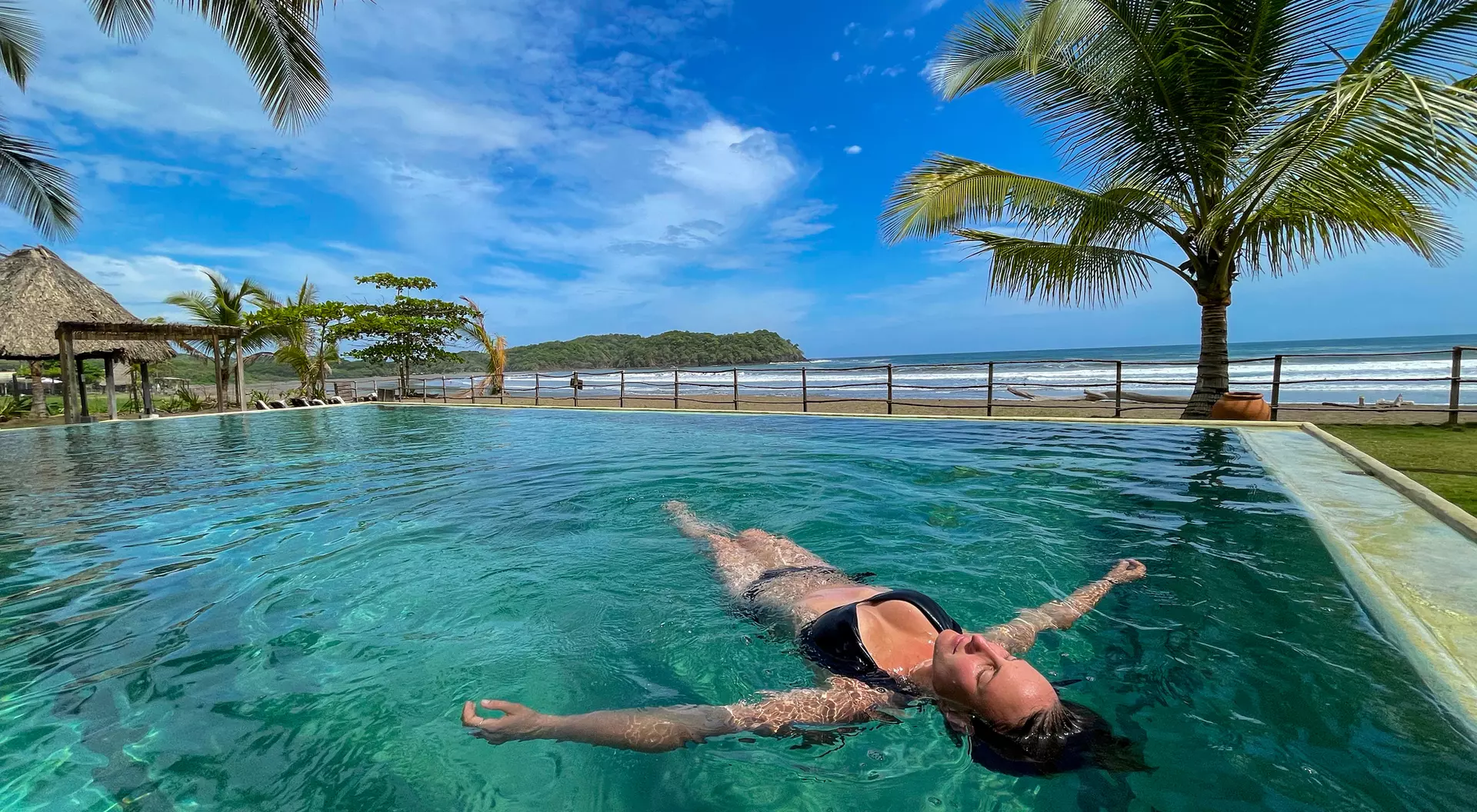 Person floating in a pool with palm trees and ocean in the background under a sunny blue sky.