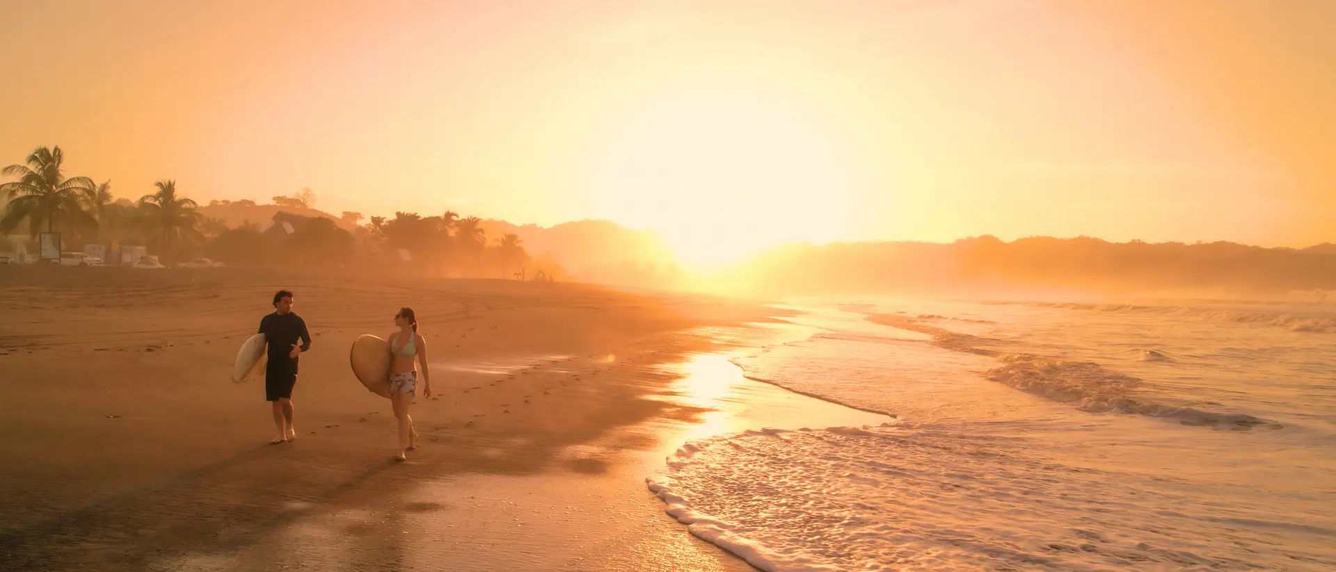 Two surfers walk along a beach at sunset, with gentle waves lapping at the shore and palm trees in the background.