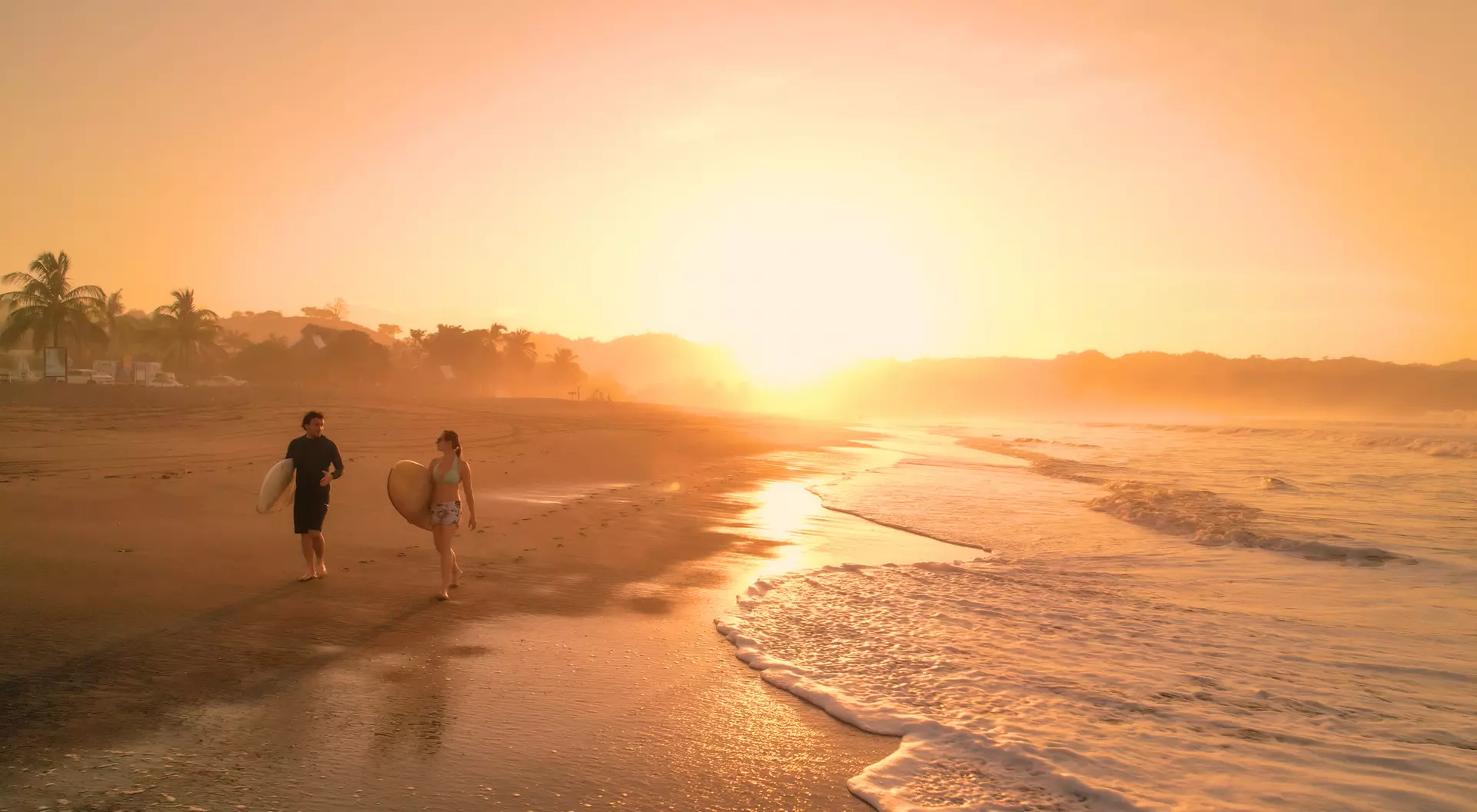 Two surfers walk along a beach at sunset, with gentle waves lapping at the shore and palm trees in the background.