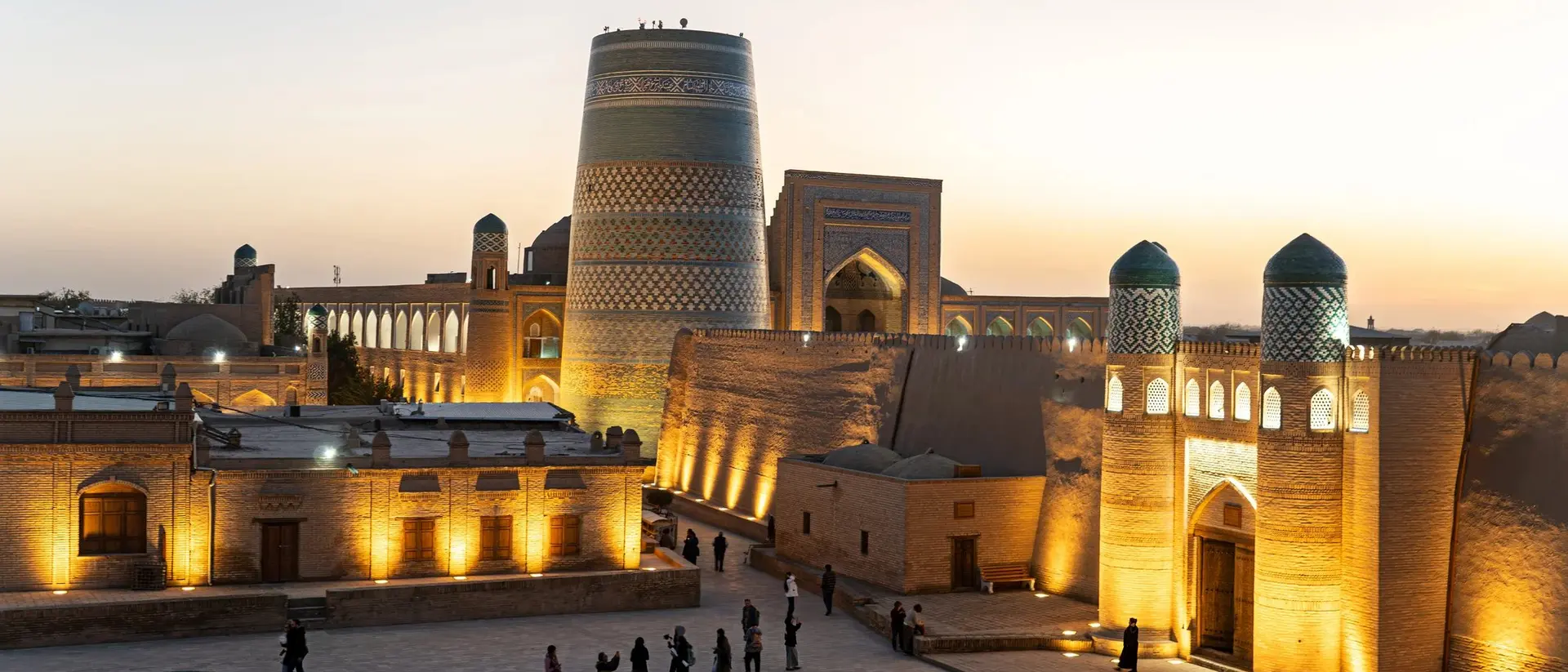 Illuminated historic buildings at sunset in a central Asian city, with people walking in a square.
