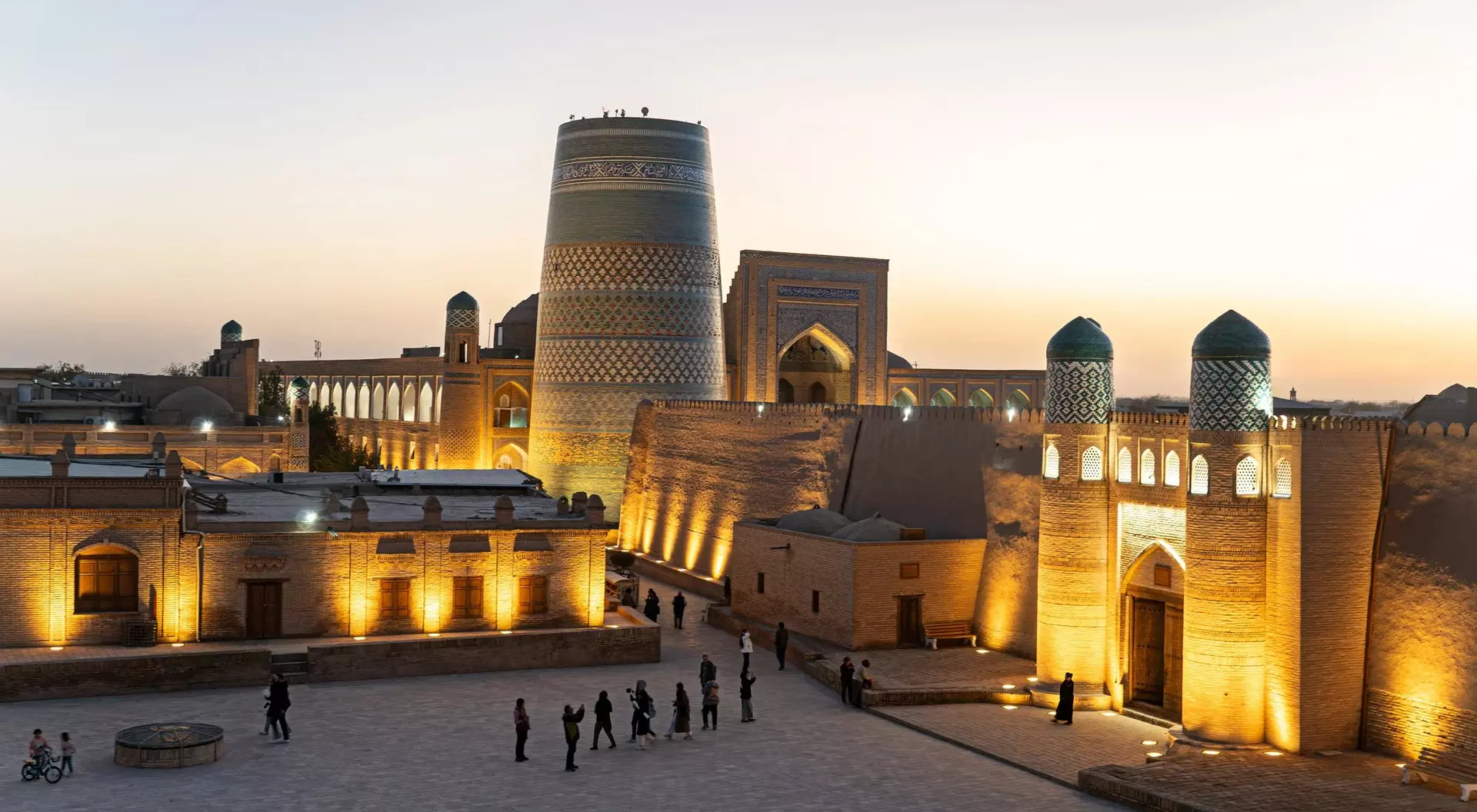 Illuminated historic buildings at sunset in a central Asian city, with people walking in a square.