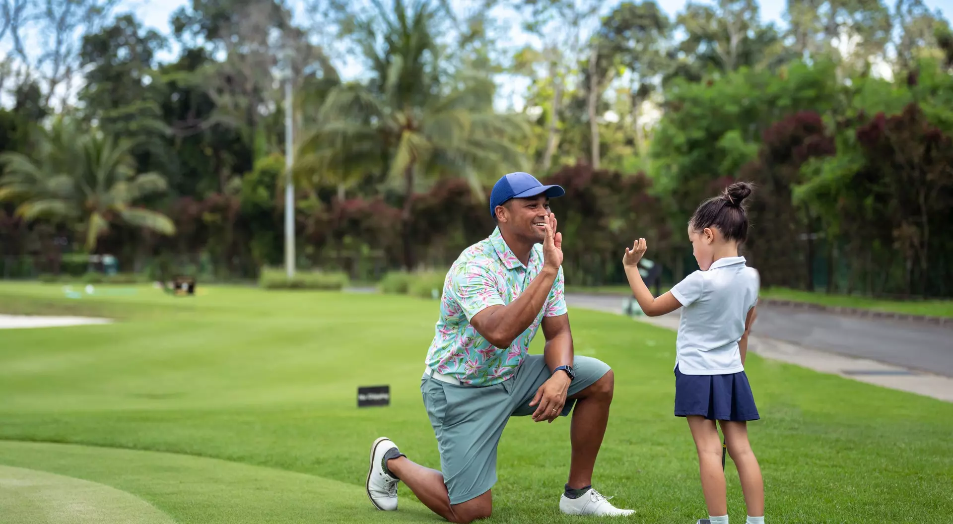 A man kneels on a golf course while a girl, wearing a golf outfit, playfully gestures with her hand.