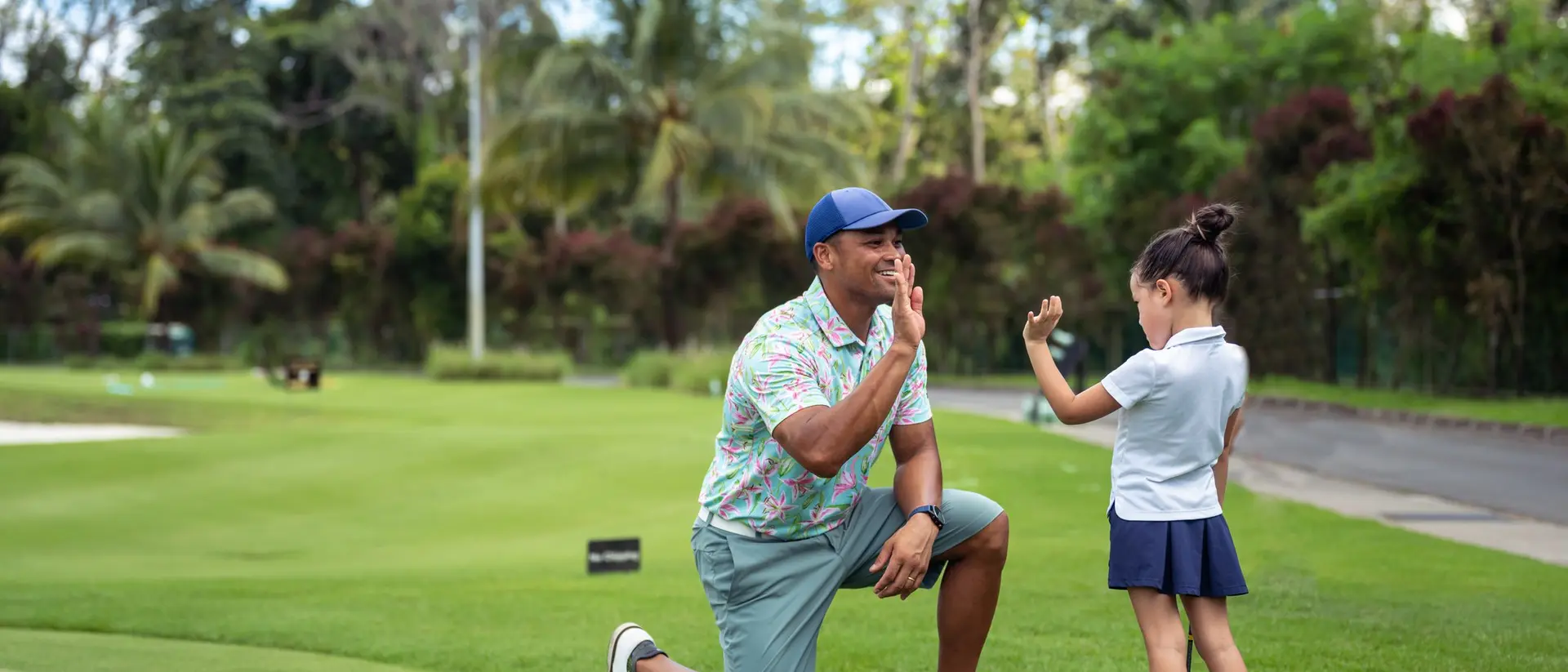 A man kneels on a golf course while a girl, wearing a golf outfit, playfully gestures with her hand.