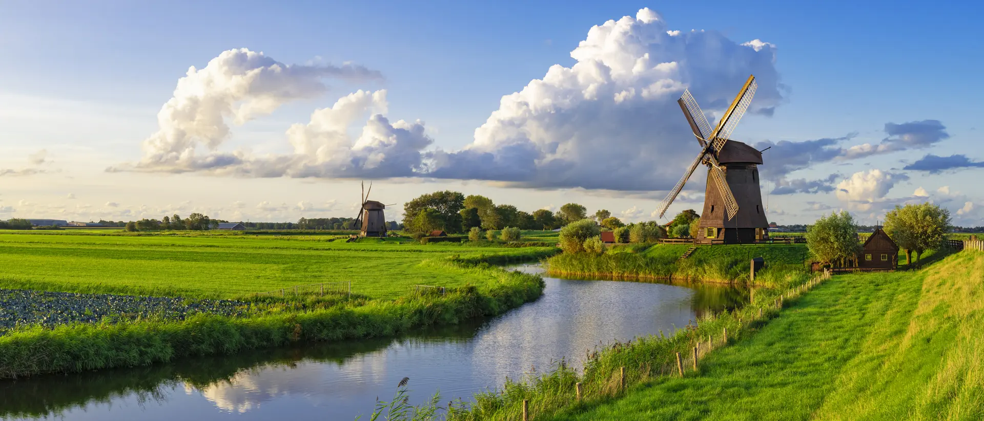 A scenic landscape featuring green fields, a river, and traditional windmills under a blue sky with fluffy clouds.