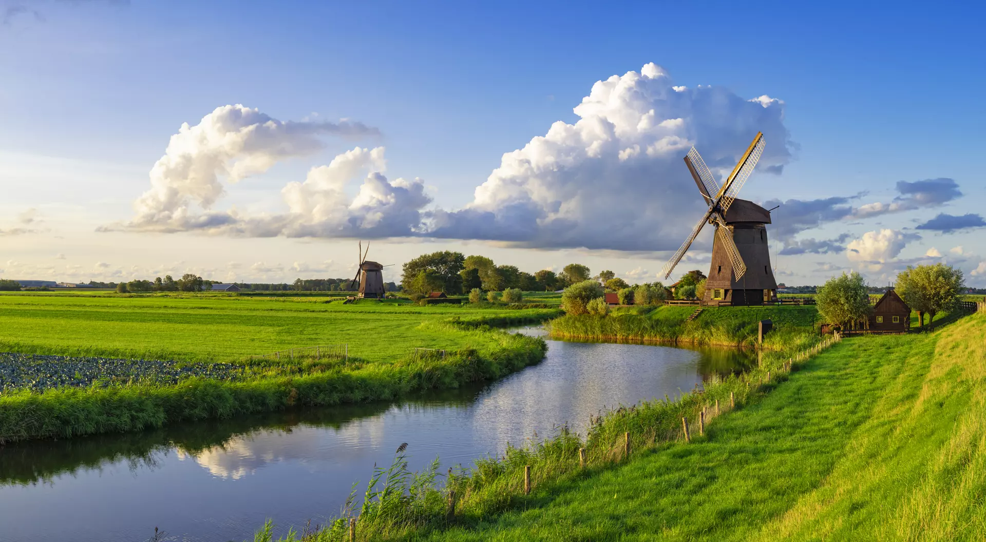 A scenic landscape featuring green fields, a river, and traditional windmills under a blue sky with fluffy clouds.