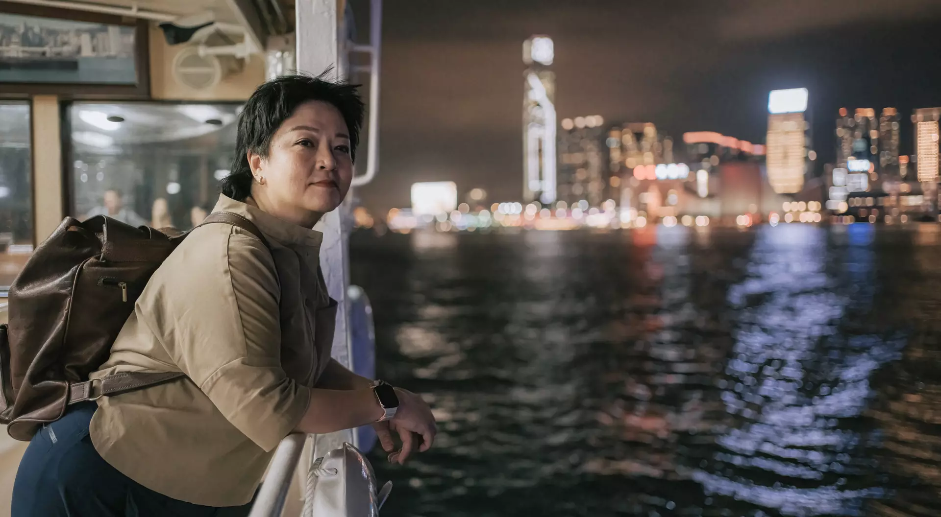 A person leans on a railing of a boat, gazing at a city skyline illuminated at night. Water ripples below.