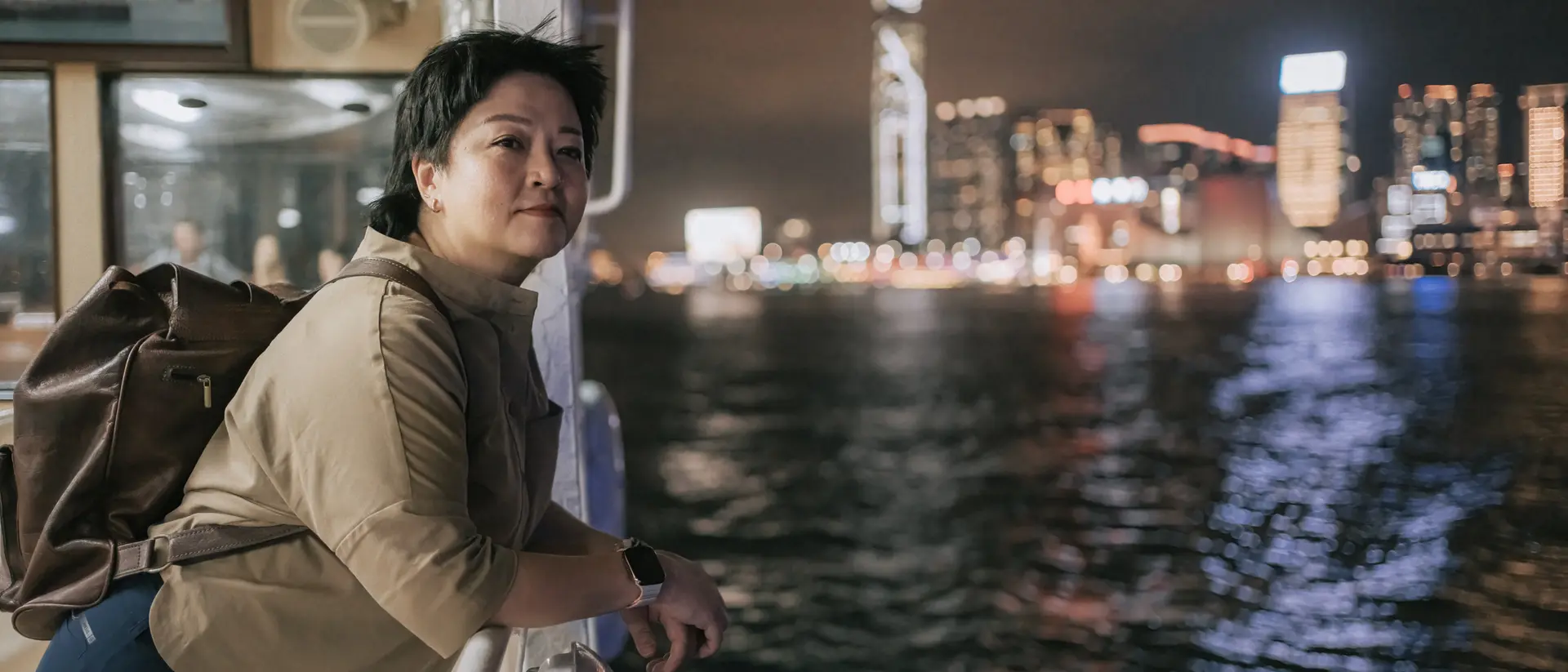 A person leans on a railing of a boat, gazing at a city skyline illuminated at night. Water ripples below.