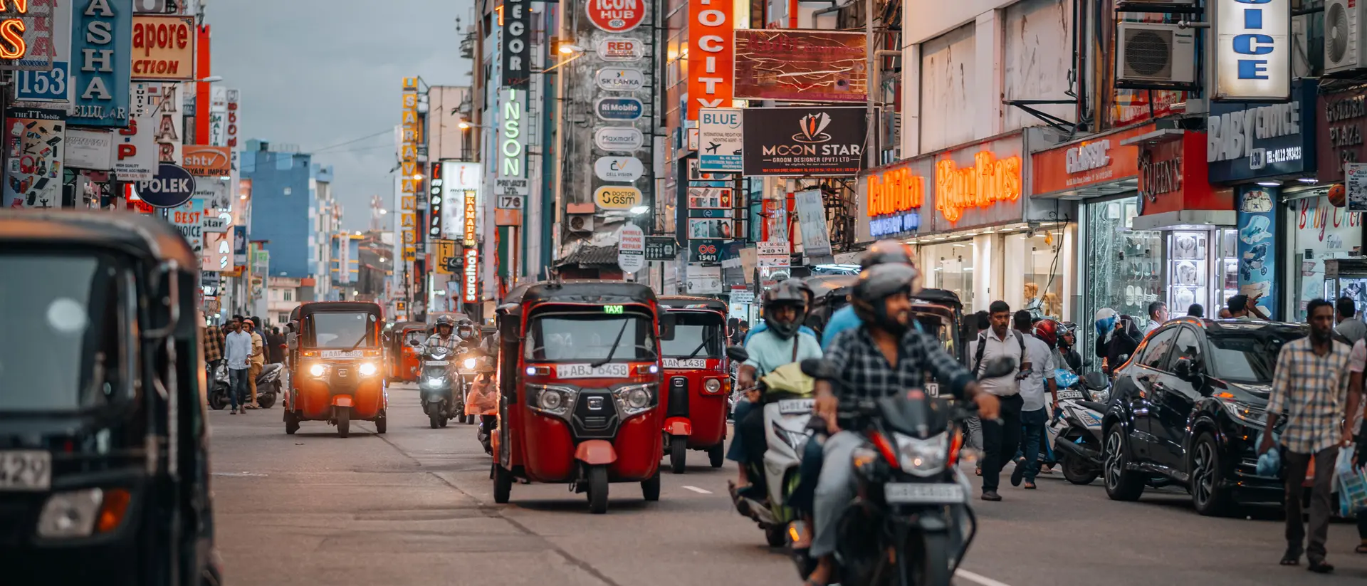 Busy street scene with tuk-tuks, motorcycles, and colorful storefronts illuminated by neon signs.