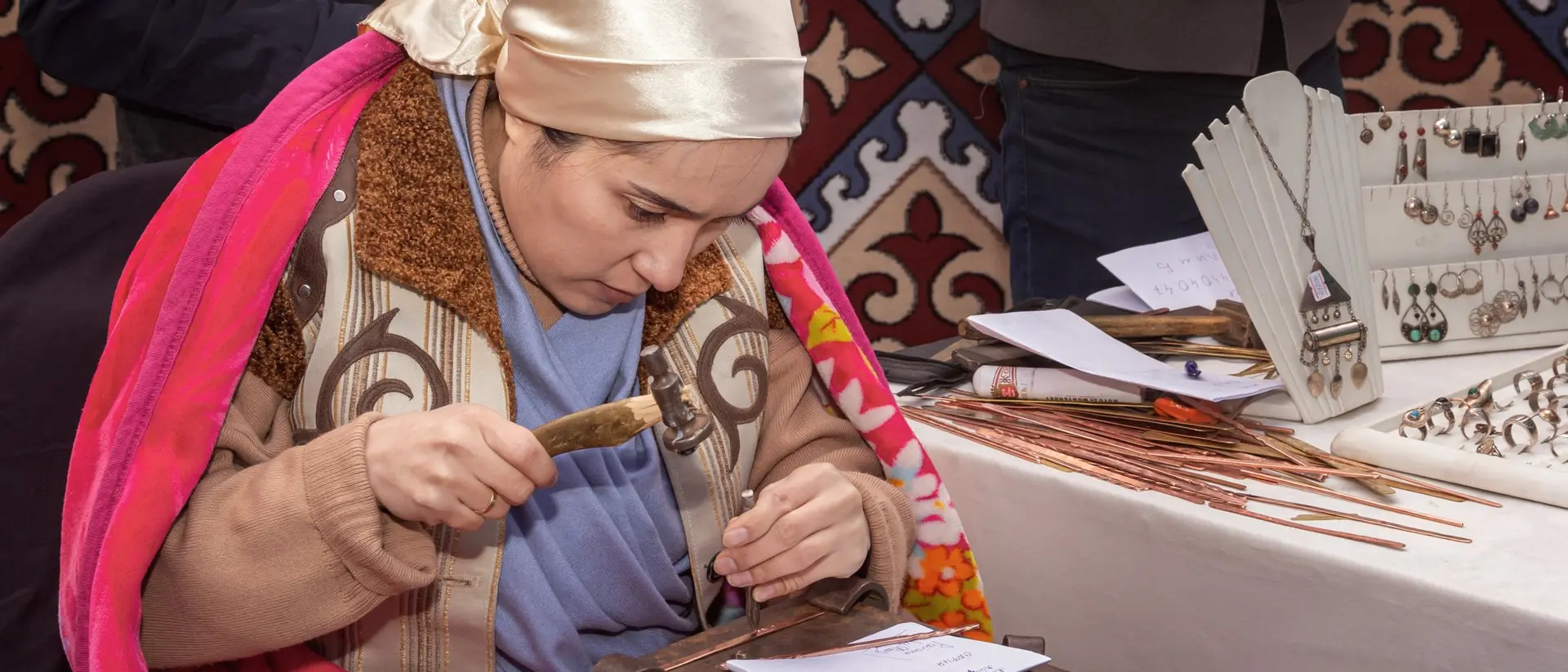 A woman in traditional attire works on a craft at a table with various tools and decorative items in the background.