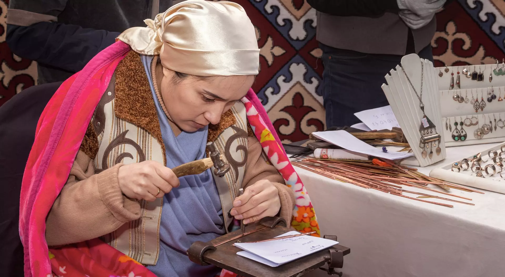 A woman in traditional attire works on a craft at a table with various tools and decorative items in the background.