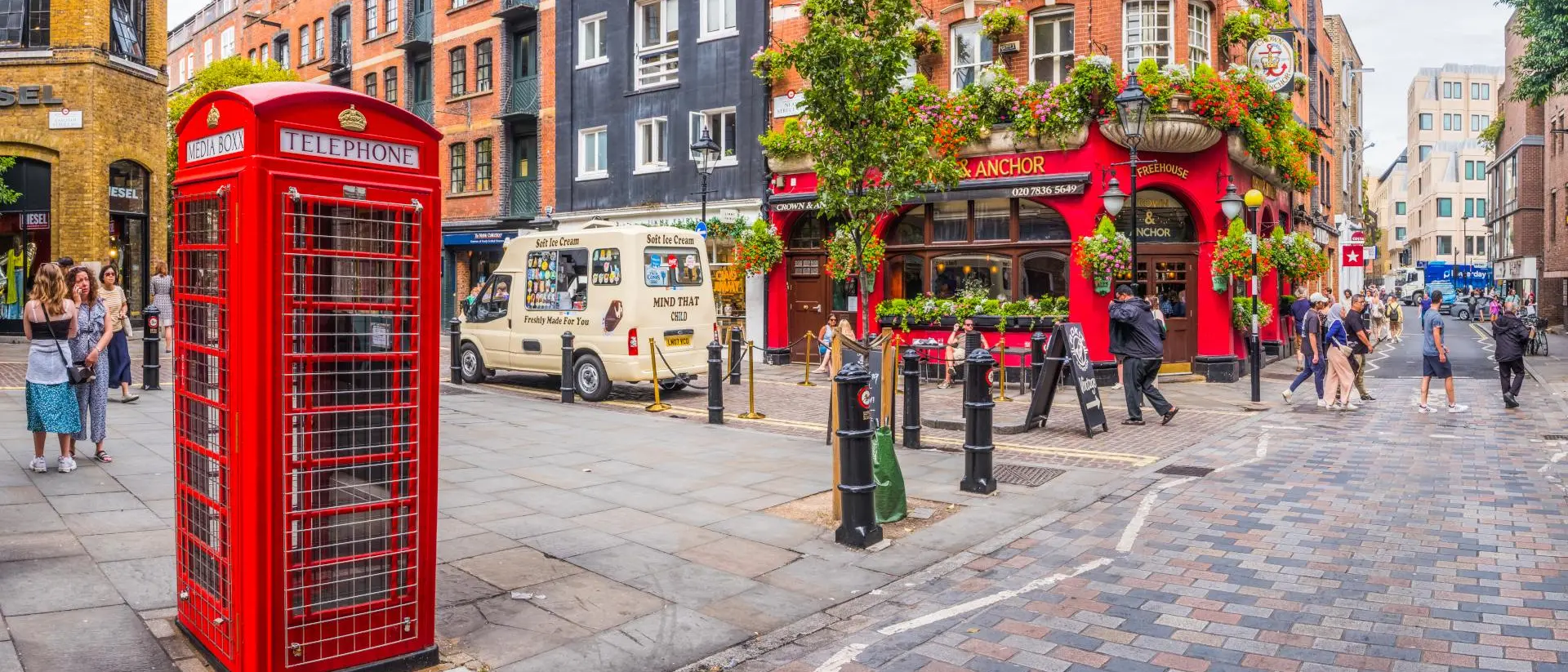 A vibrant street scene featuring a red telephone box, a pub adorned with flowers, and people enjoying the urban atmosphere.