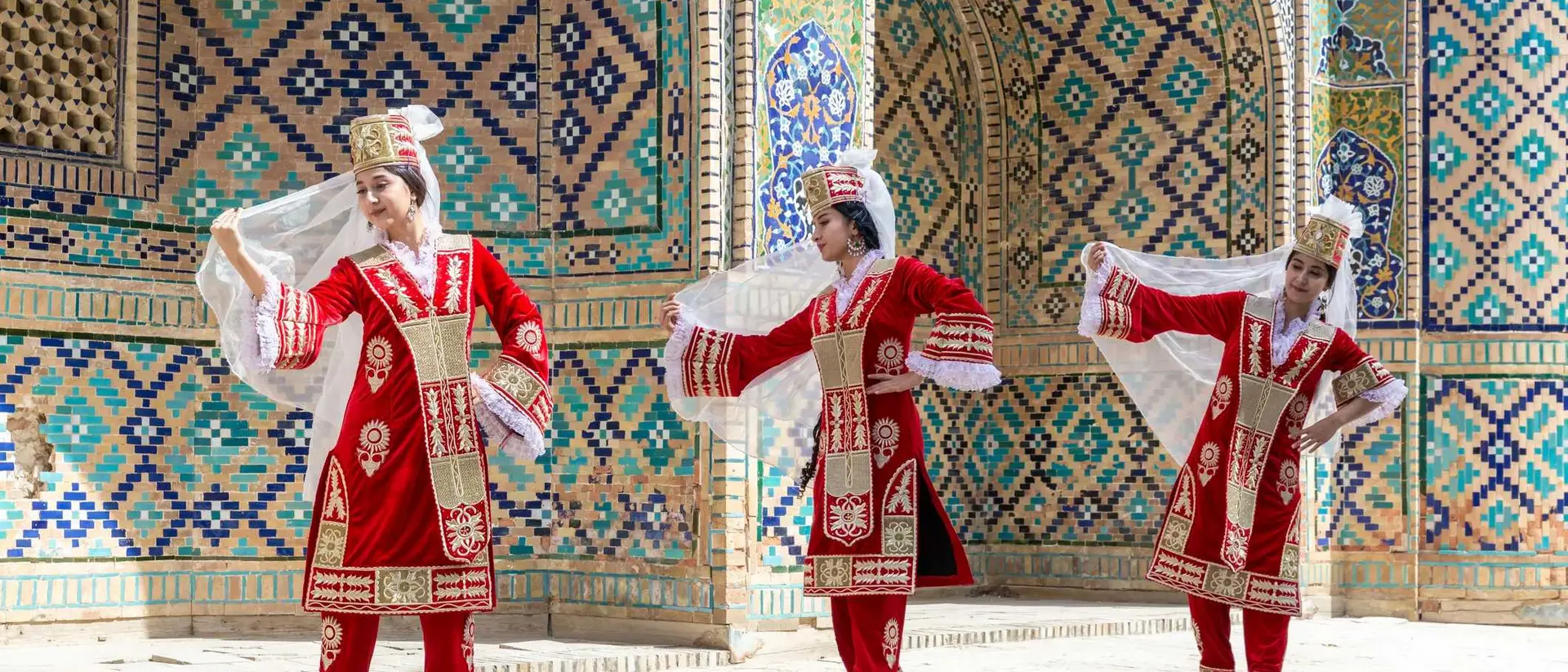 Three dancers in traditional red costumes perform against a colorful, tiled backdrop. They hold flowing white veils.