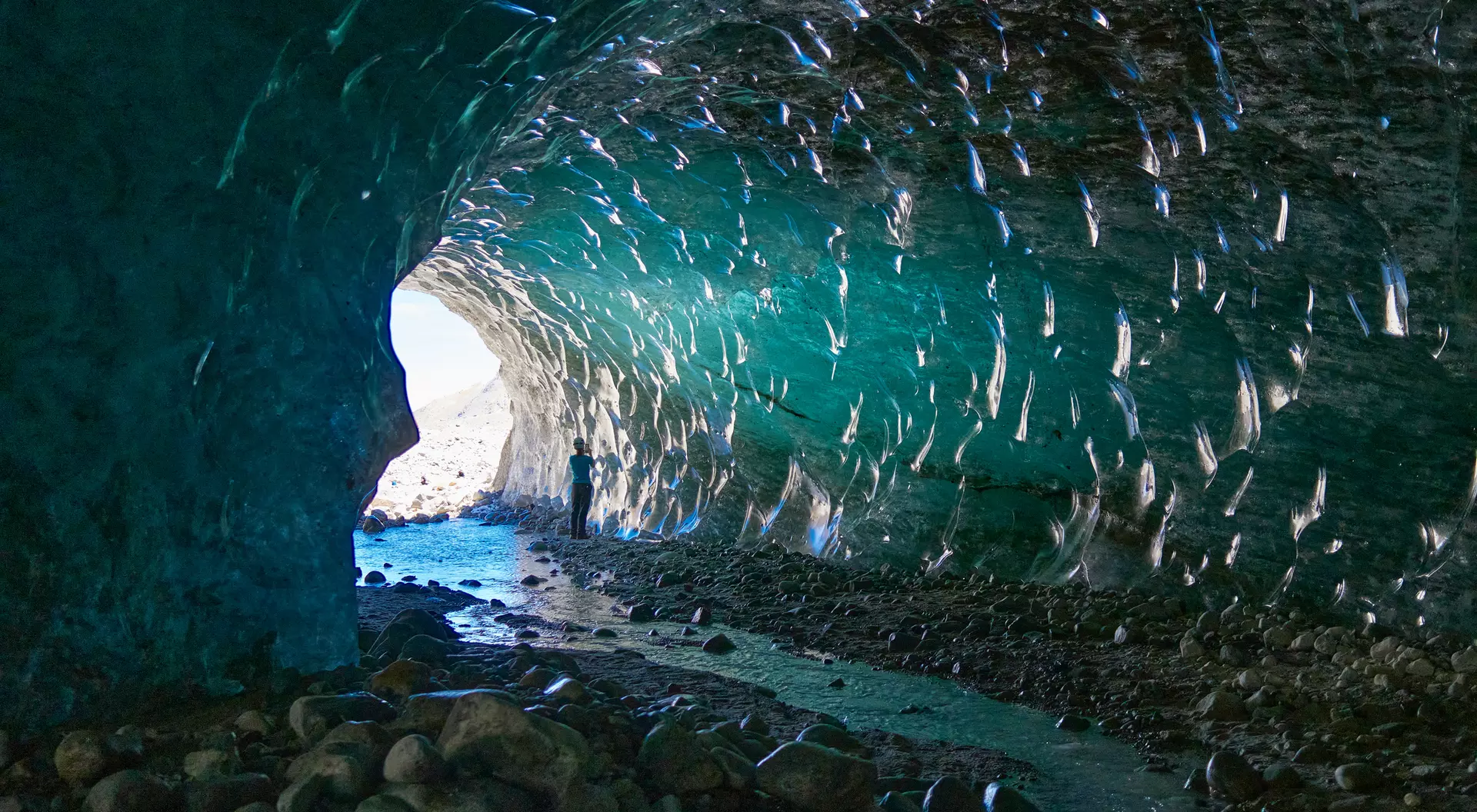 Person standing inside a glacial cave, with shimmering ice and water reflecting light from the entrance.