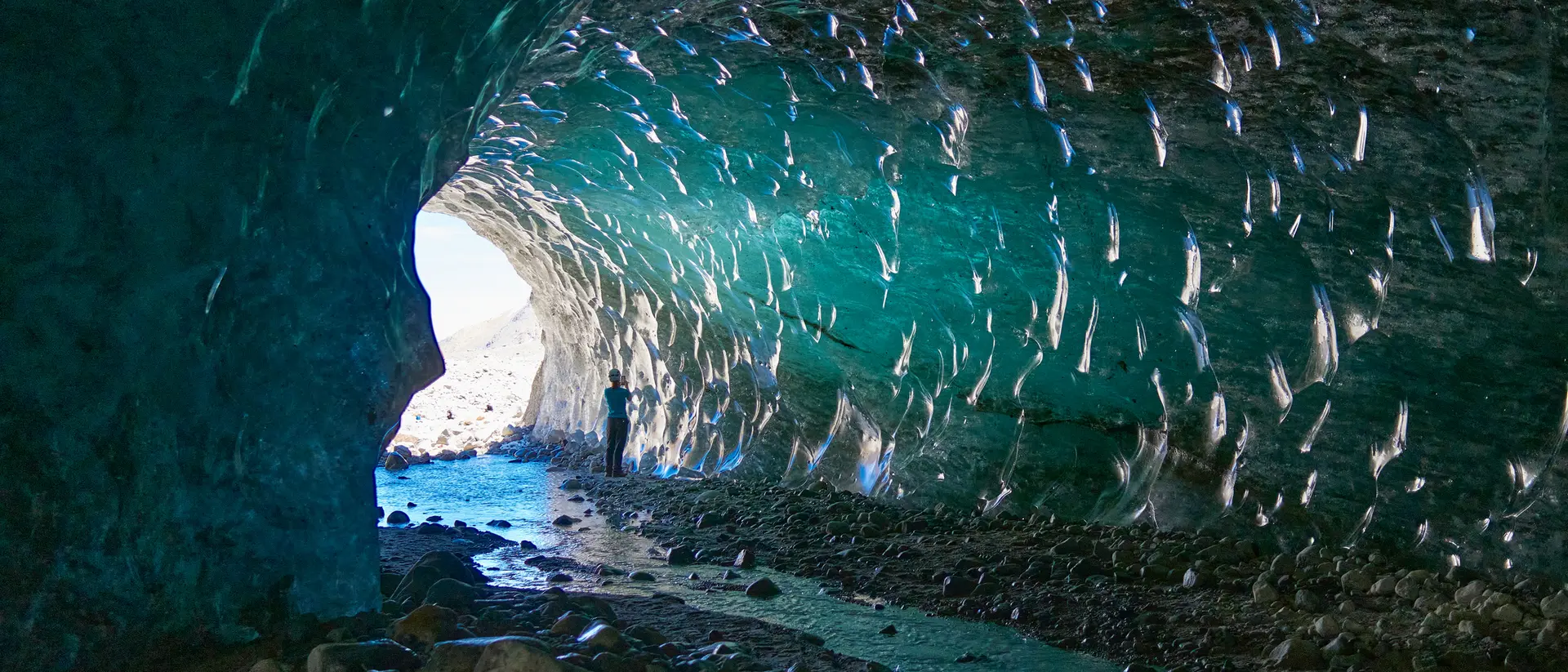 Person standing inside a glacial cave, with shimmering ice and water reflecting light from the entrance.