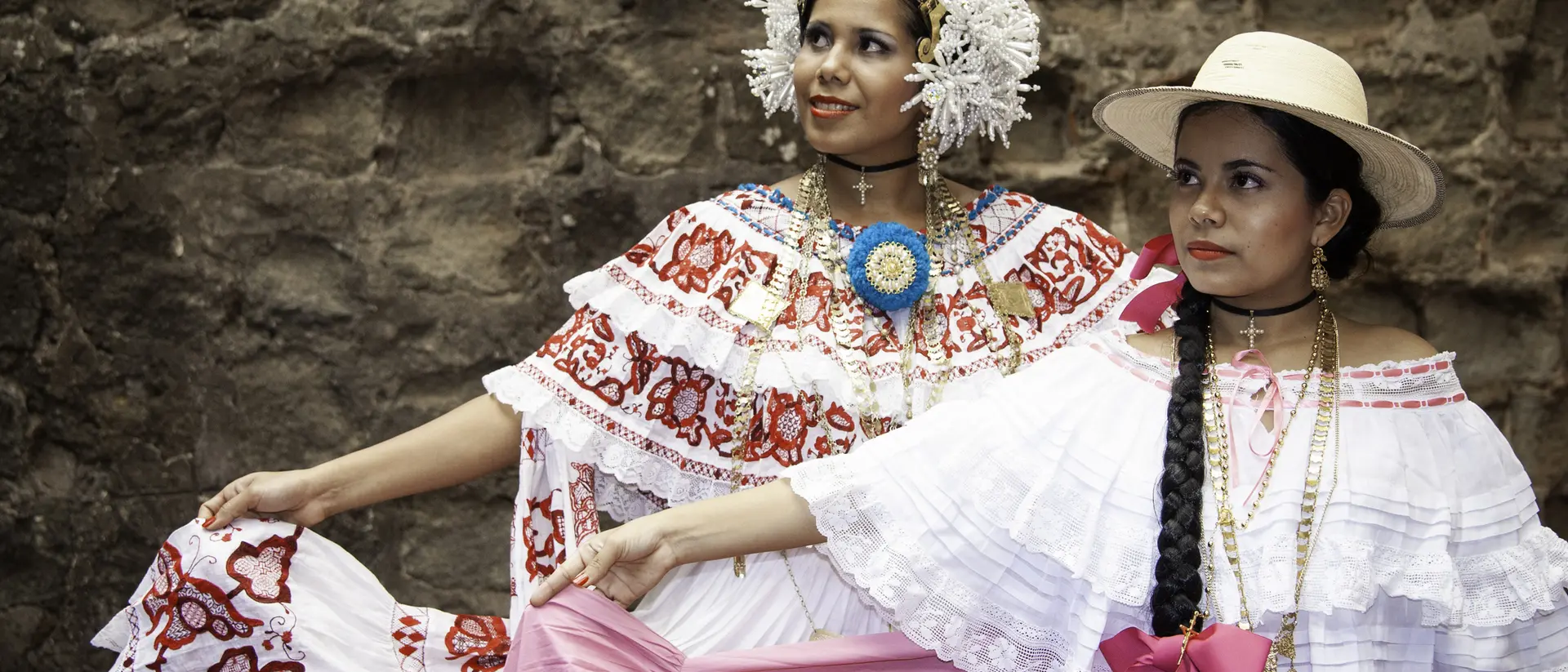 Two women in vibrant traditional dresses pose against a stone wall, showcasing colorful patterns and cultural accessories.
