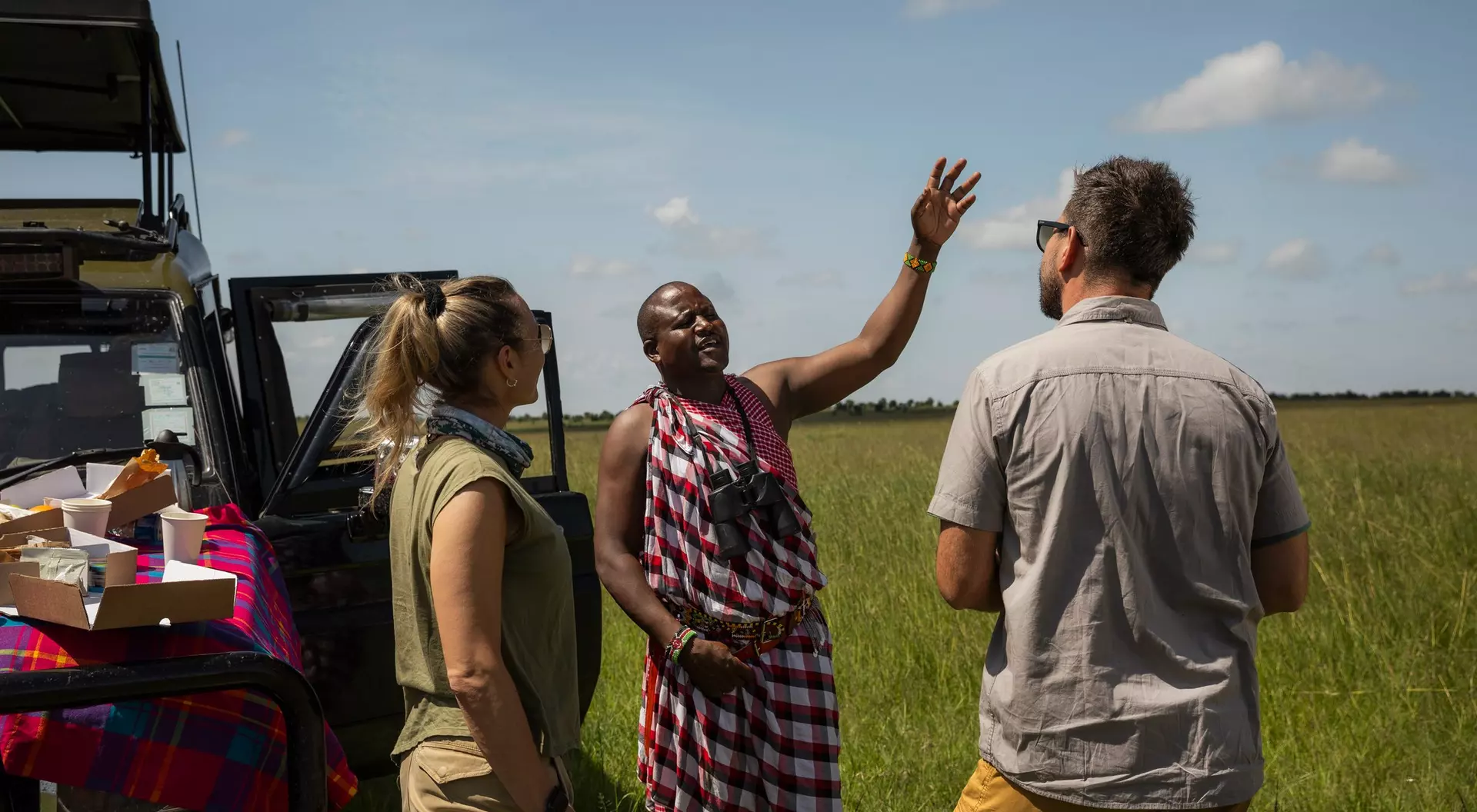 Three people engage in conversation outdoors near a vehicle, surrounded by a grassy landscape under a blue sky.