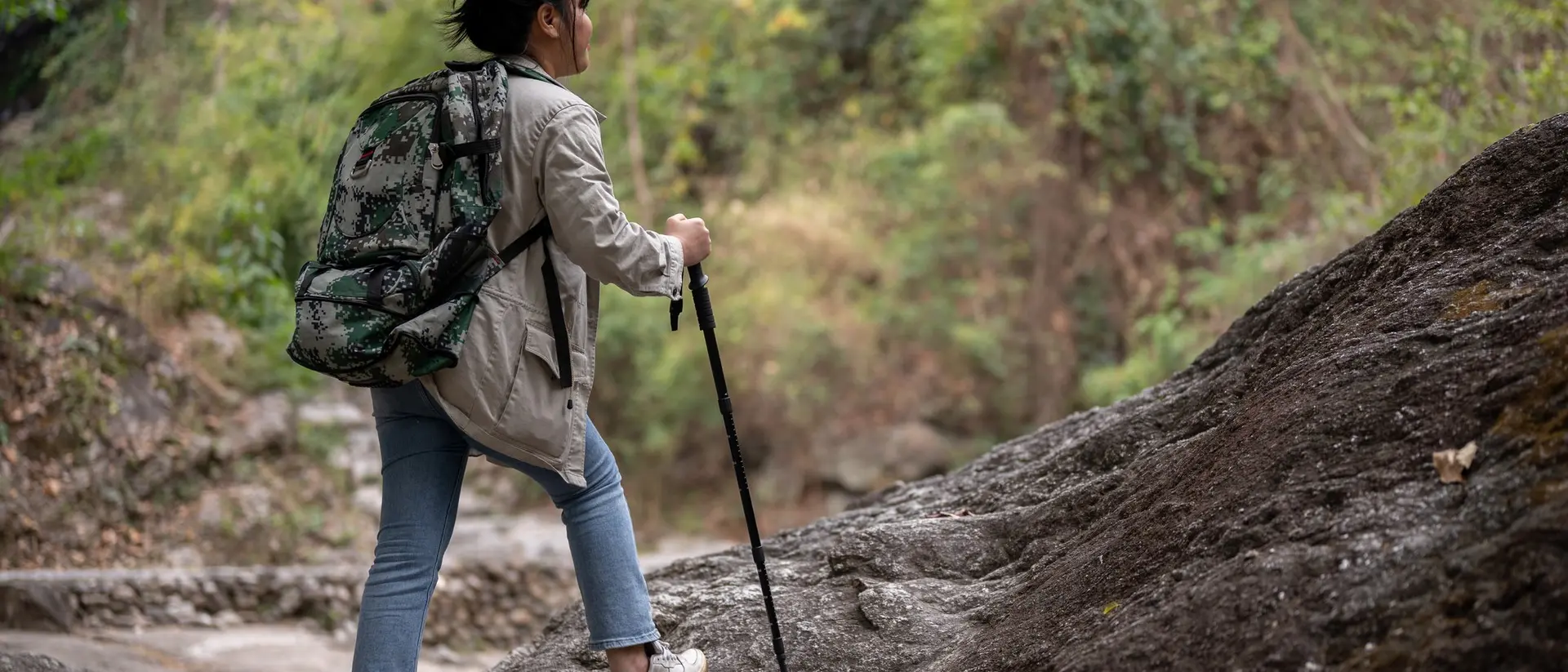 A person hiking up a rocky path, carrying a large backpack and using a walking stick, surrounded by greenery.
